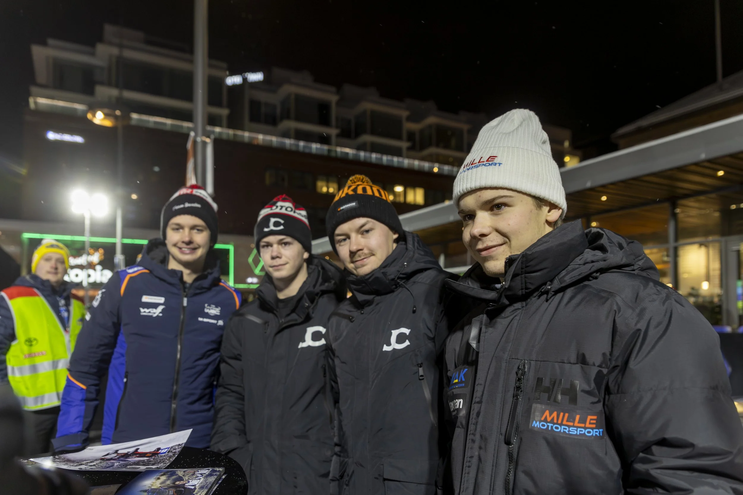 A group of young men wearing racing jackets and beanies, standing outdoors at night, in an urban setting with buildings and bright lights.