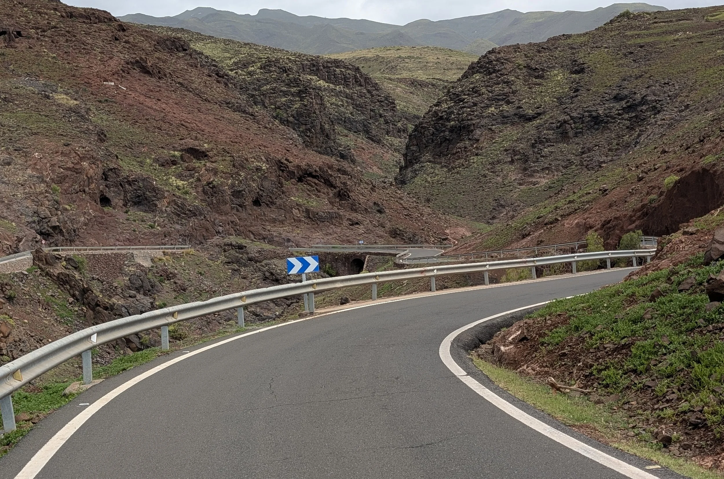 A winding mountain road with guardrails, surrounded by rocky, green-covered hills and mountains.