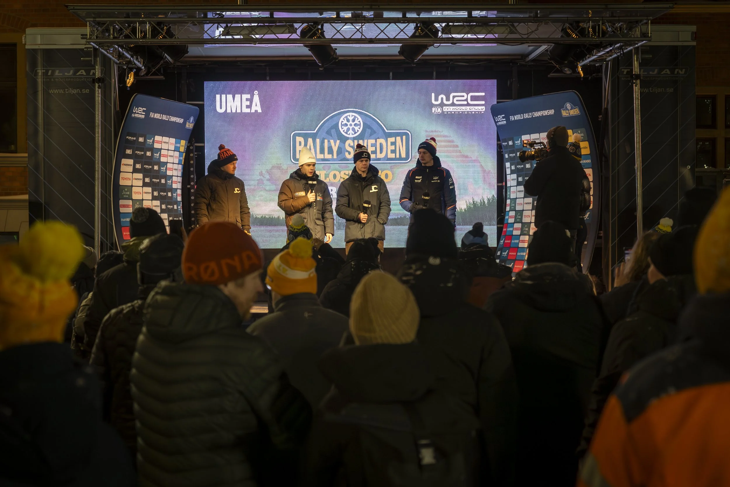 Four rally race drivers dressed in winter coats and hats standing on stage at a rally Sweden event, with a crowd in winter clothing watching in the foreground. A large screen behind them displays the rally Sweden logo, sponsors, and event information