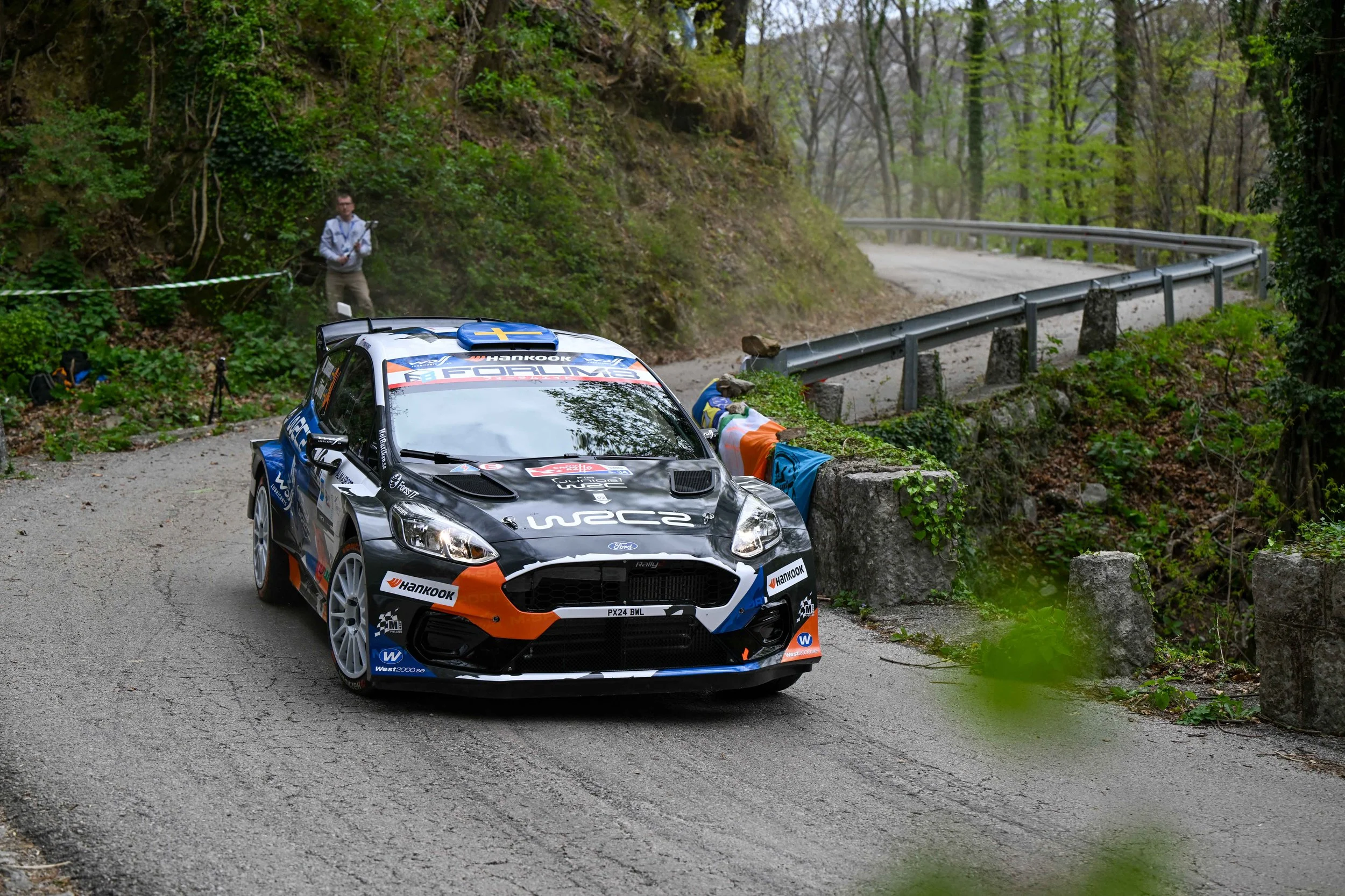 A rally car on a narrow, winding mountain road surrounded by lush green trees. A man stands off to the side near a rope barrier, watching the car. The car is decorated with sponsor logos and racing decals.