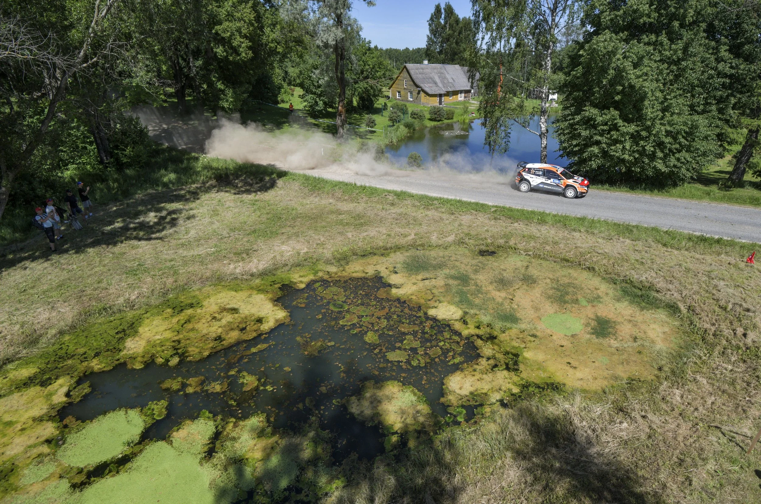 A rally car speeds along a dirt road next to a pond with green algae, dust rising behind it, surrounded by trees and a few spectators.