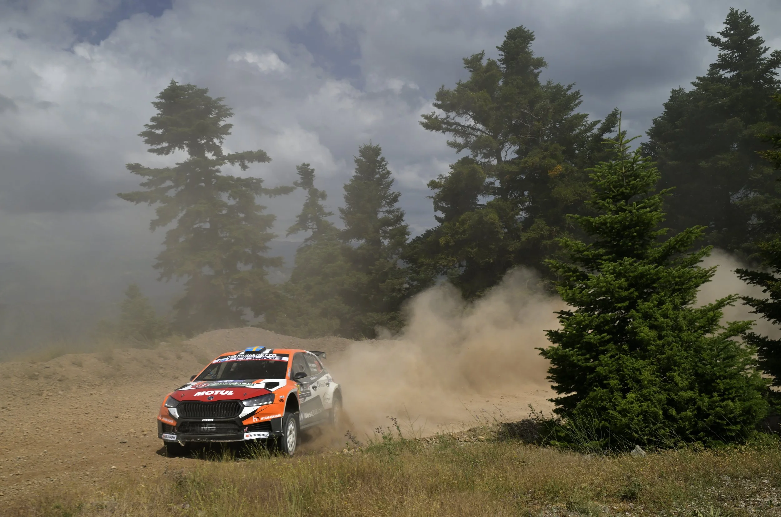 An orange and white rally car speeding on a dirt track surrounded by trees and dust clouds.