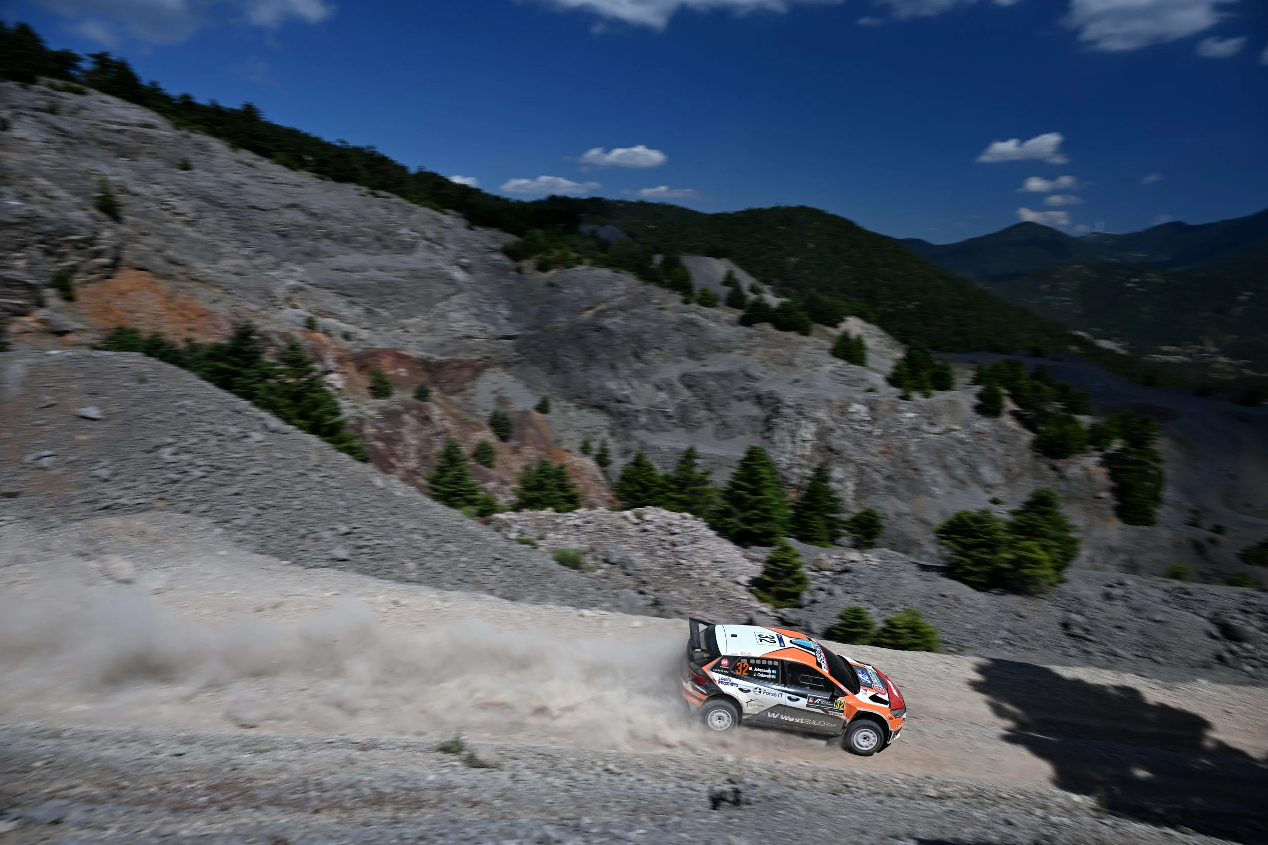 A rally car speeding on a dirt mountain road with dust trailing behind, surrounded by rugged terrain, pine trees, and distant mountains under a partly cloudy blue sky.