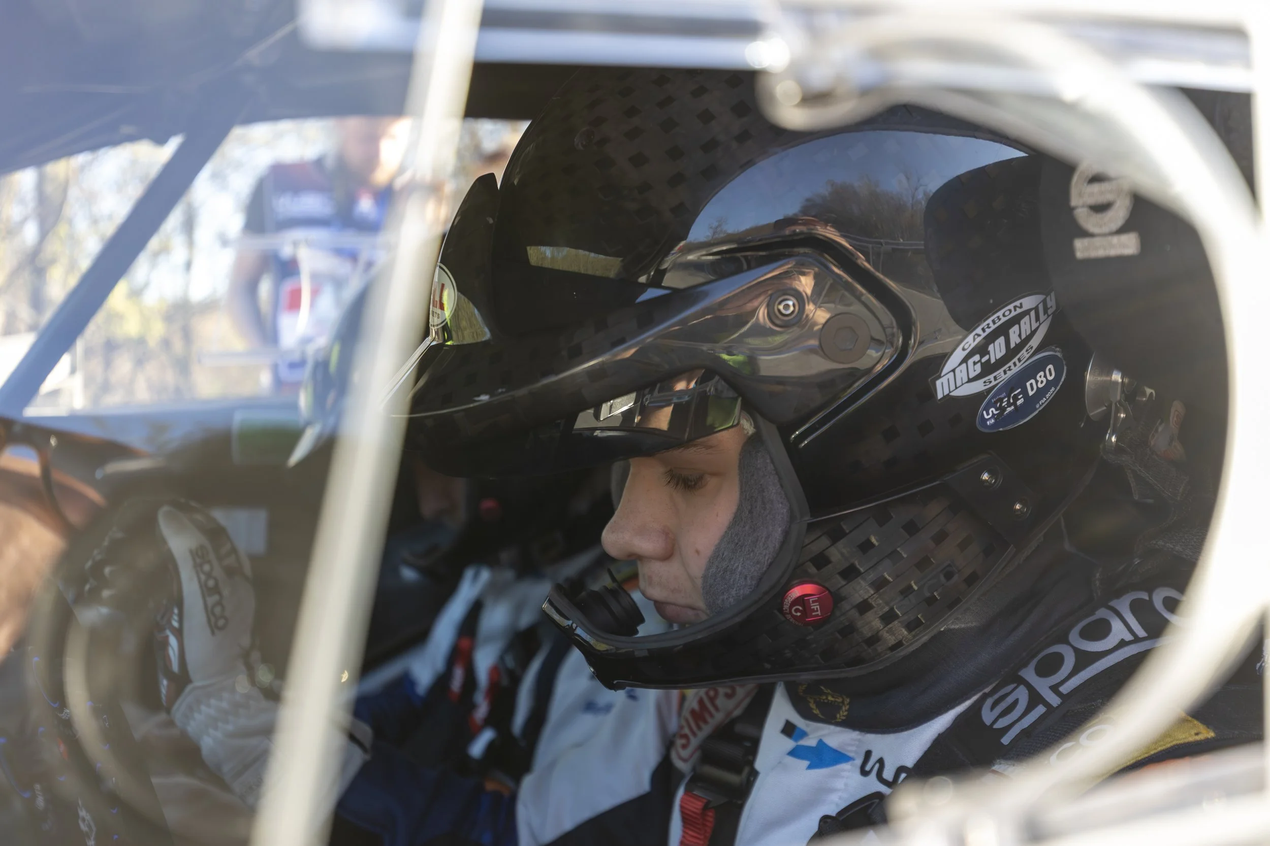 Close-up of a race car driver wearing a black carbon fiber helmet with various spots and a sticker that reads 'MAG-10 RALLY SERIES' and 'D80', with the driver’s face partially visible and focused inward.