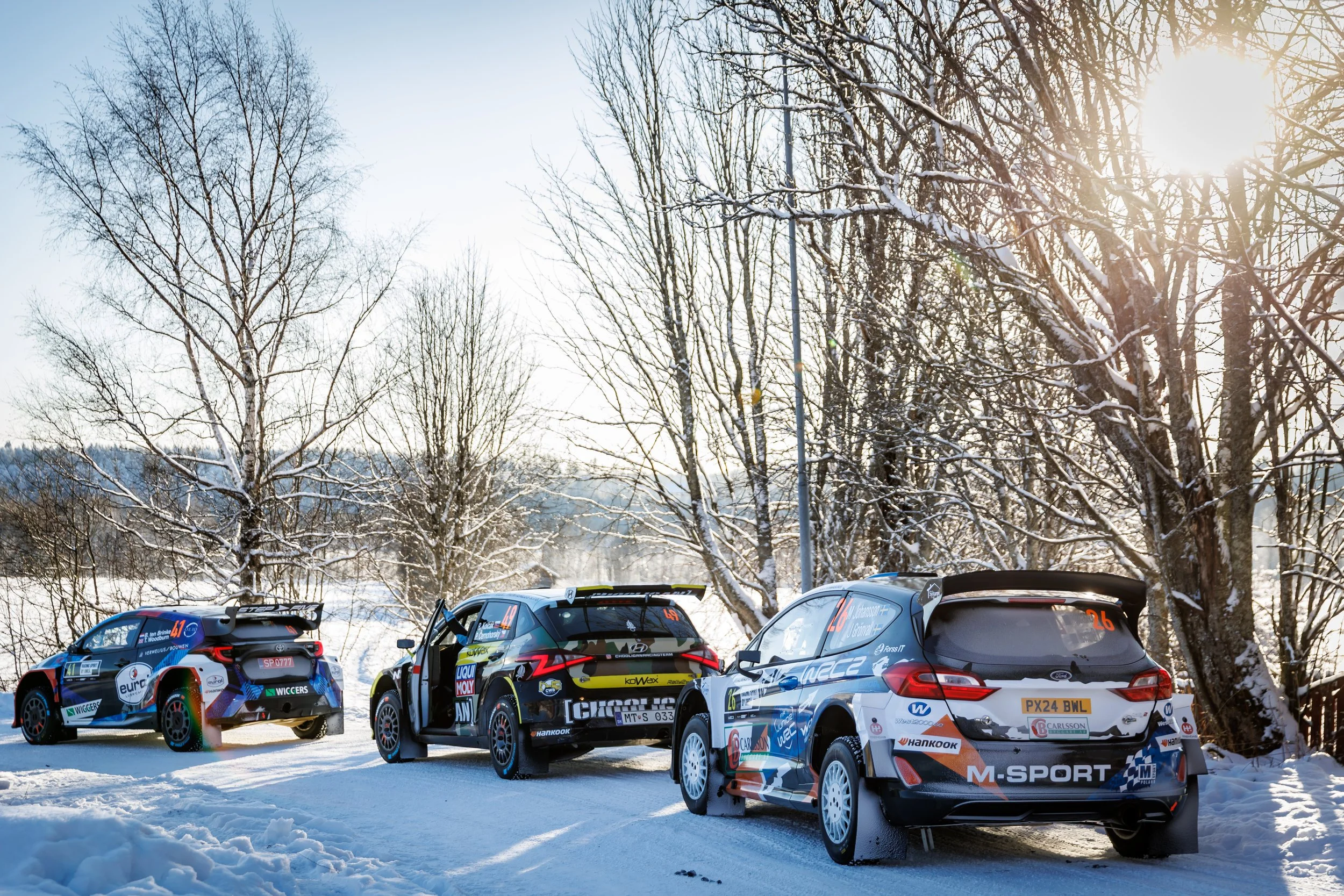 A group of race cars parked on snowy ground beneath leafless trees, with sunlight filtering through the branches.