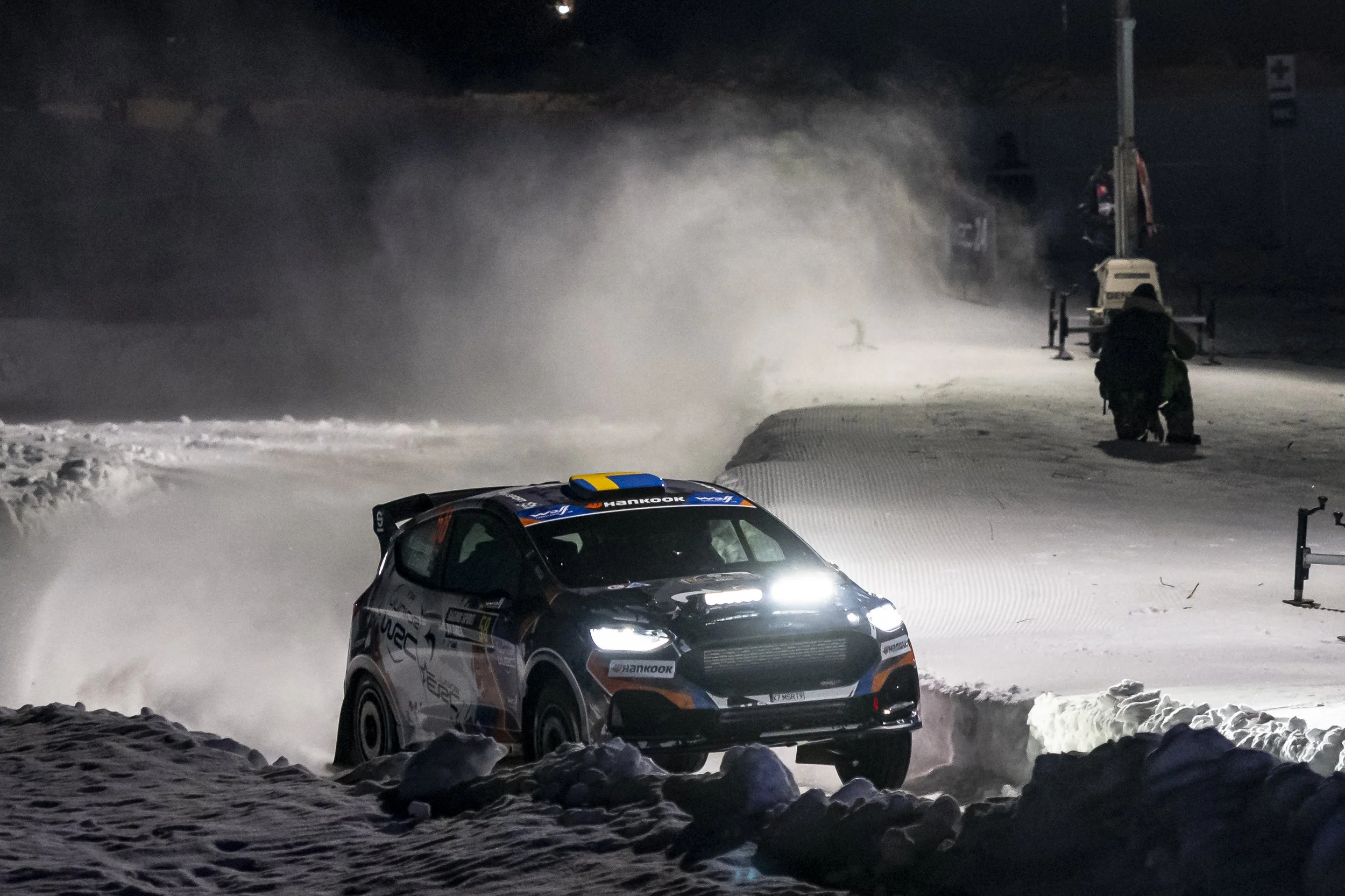 A race car speeding on a snow-covered track at night, with snow being kicked up by the tires. There is a person kneeling near the side of the track, and snow grooming equipment and signs can be seen in the background.