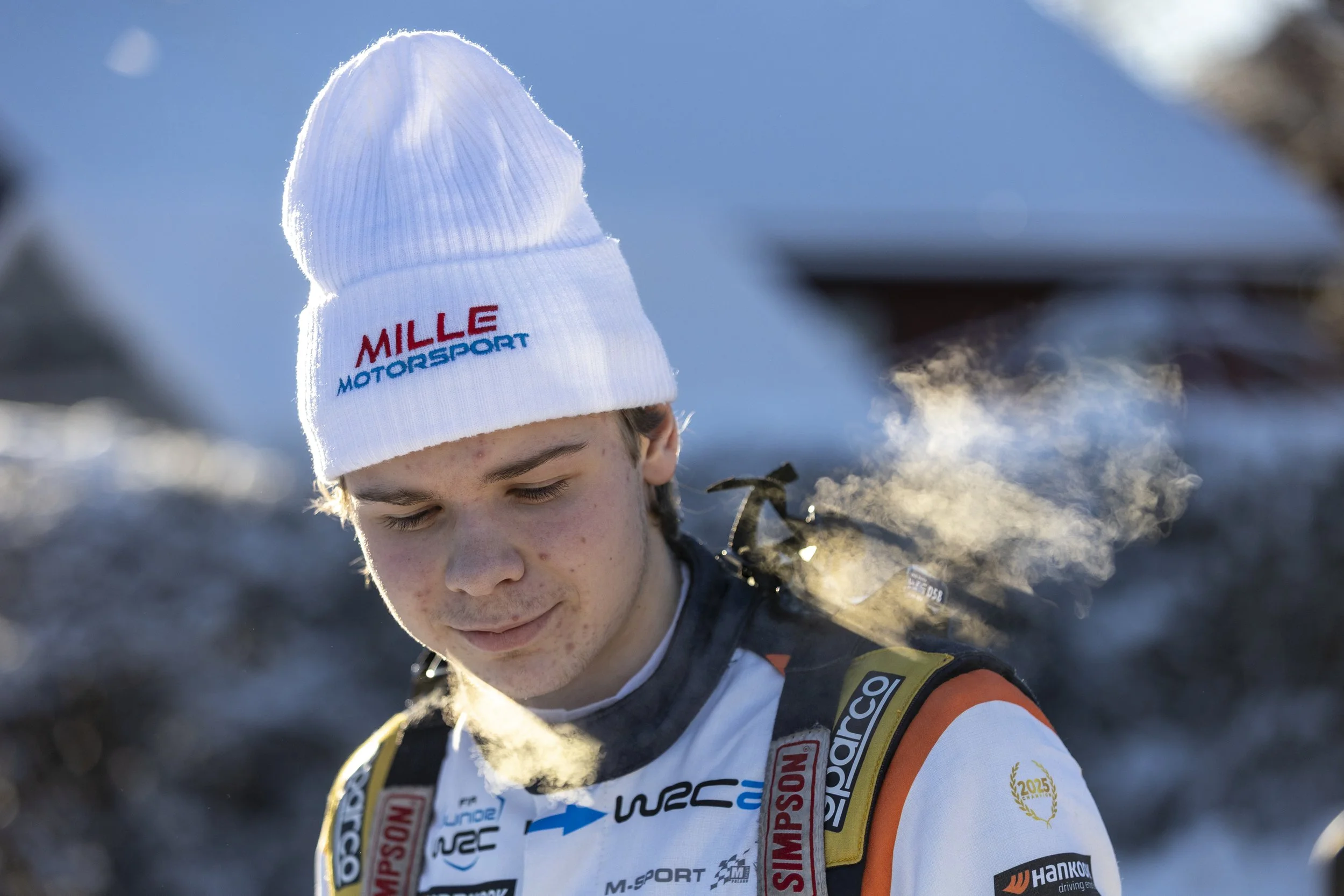 A young man in racing gear wearing a white beanie with "MILLE MOTORSPORT" embroidered on it, standing outdoors in a snowy landscape.