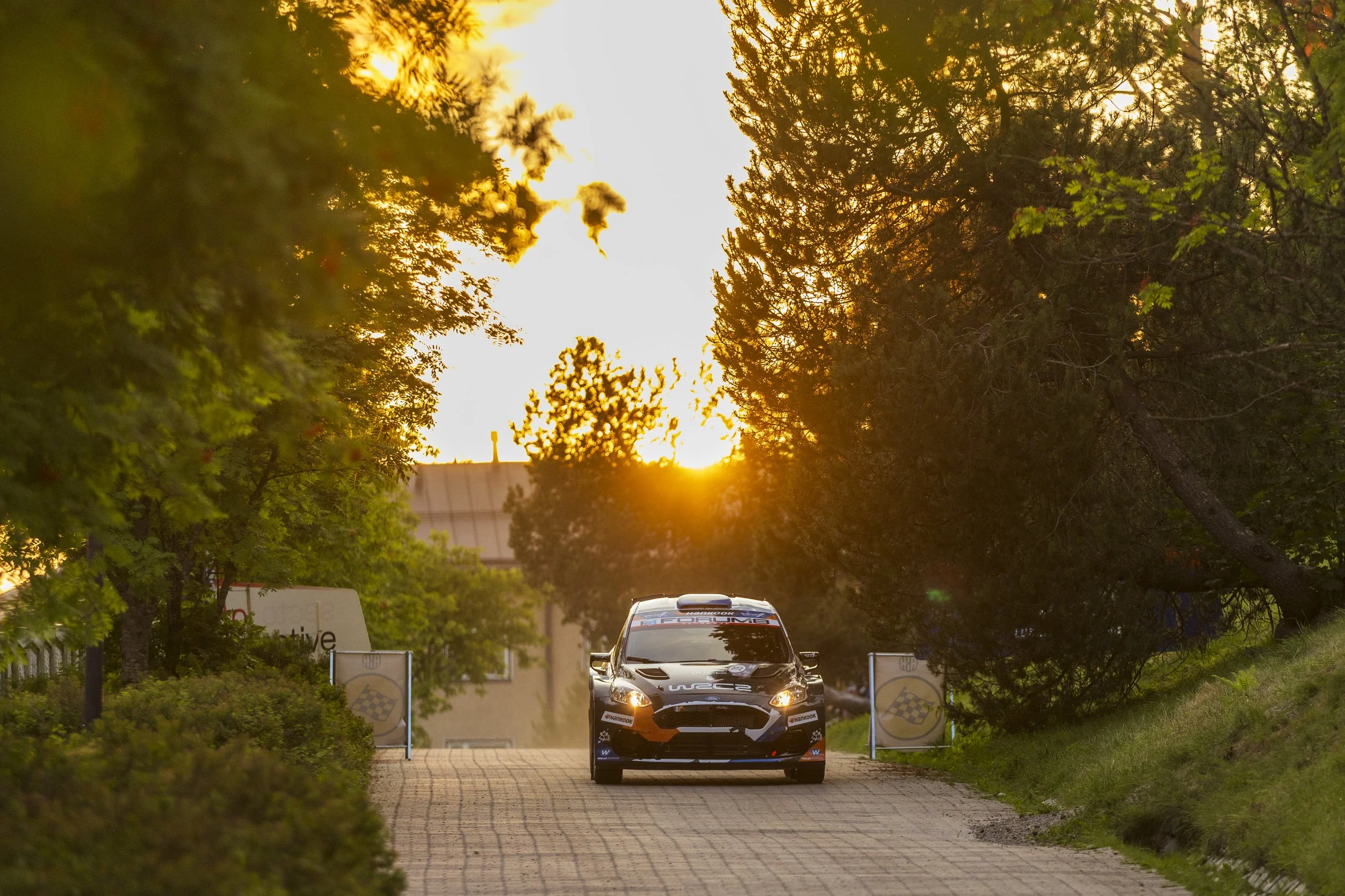A rally car driving on a road in a park during sunset, with banners and trees on each side.