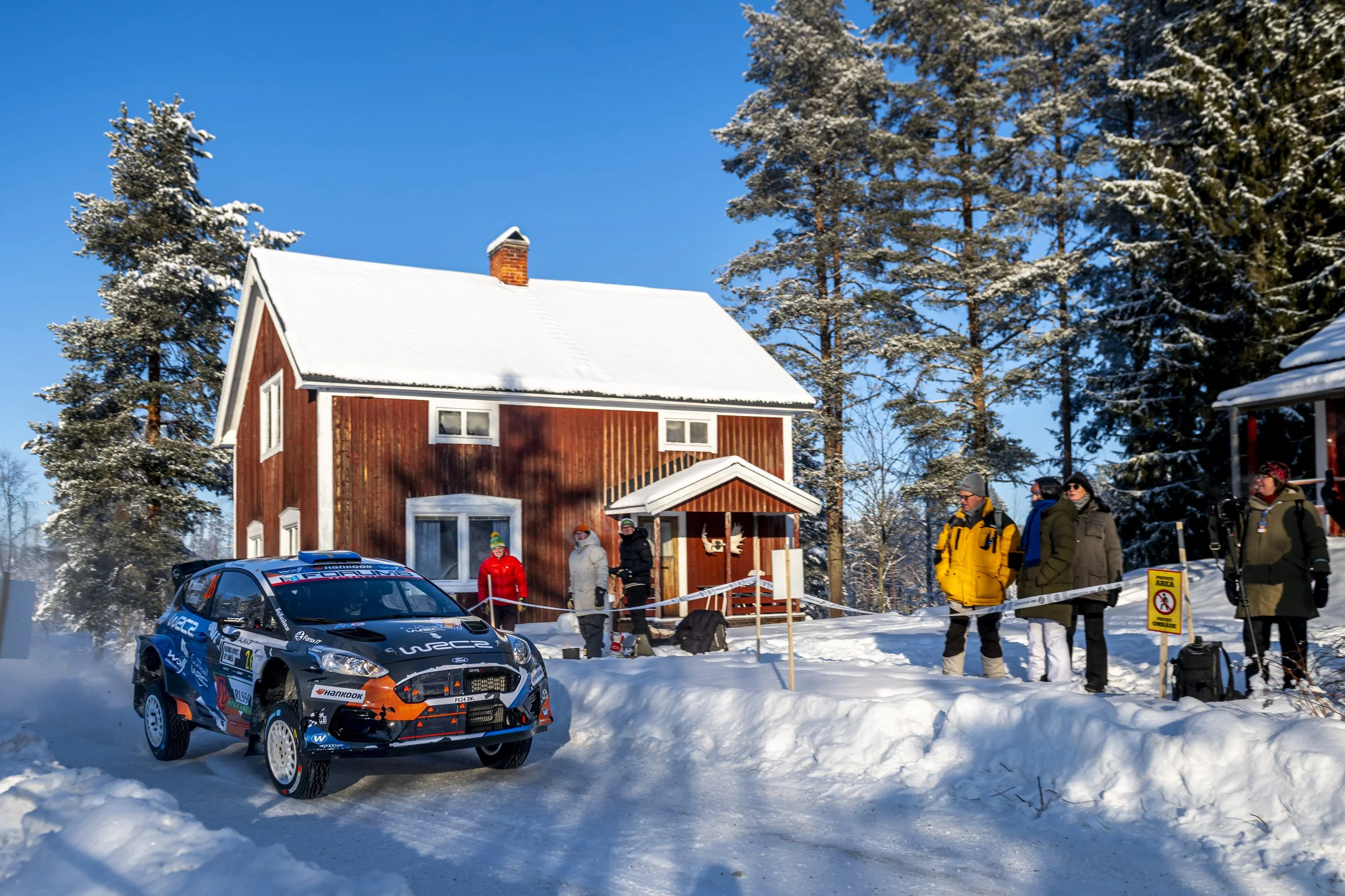 A rally car racing on snowy terrain with a red house and evergreen trees in the background, and spectators watching.