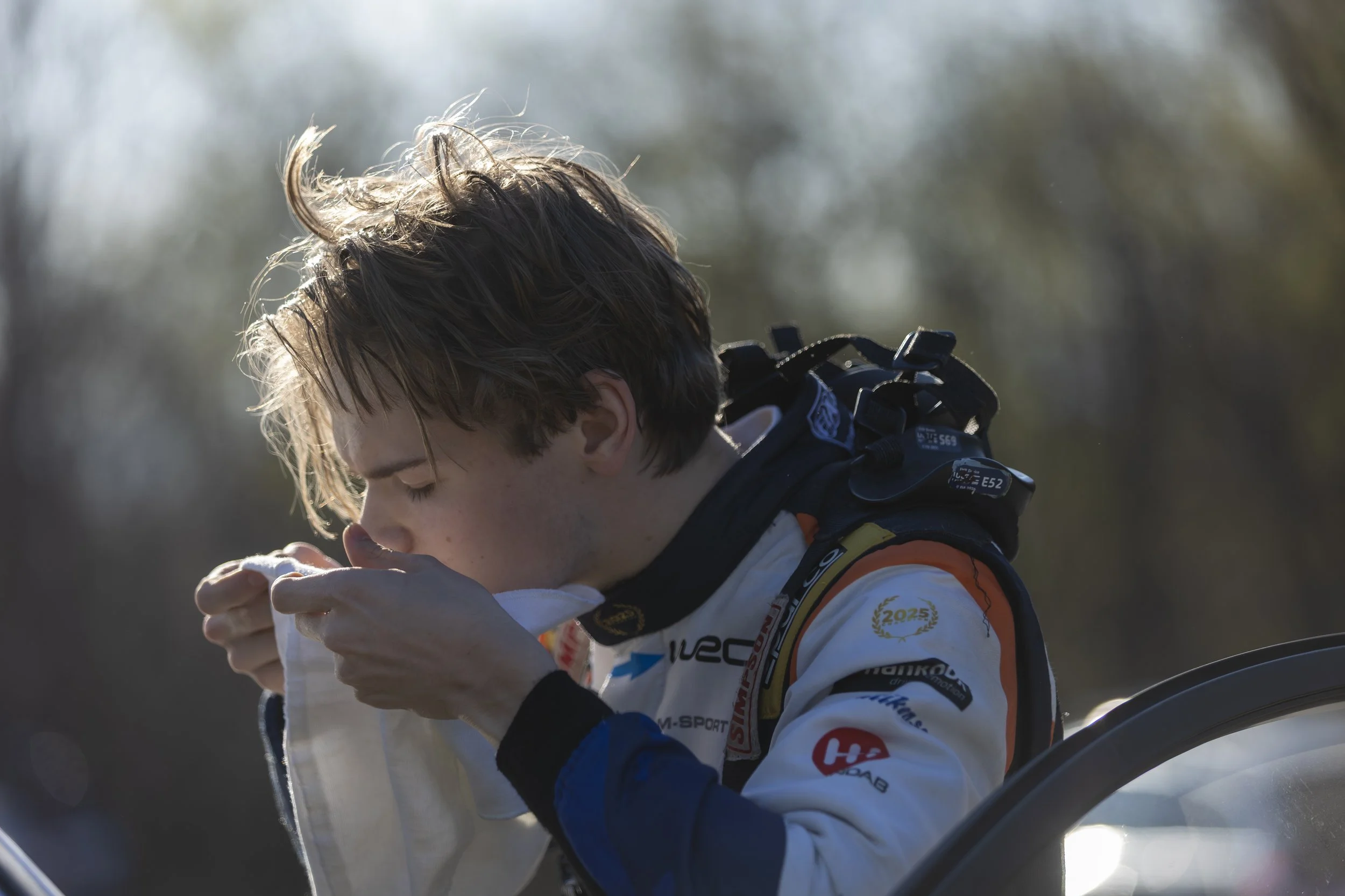 A young male race car driver with messy light brown hair, wearing a white racing suit with various sponsor logos, is smelling or tasting a small object.