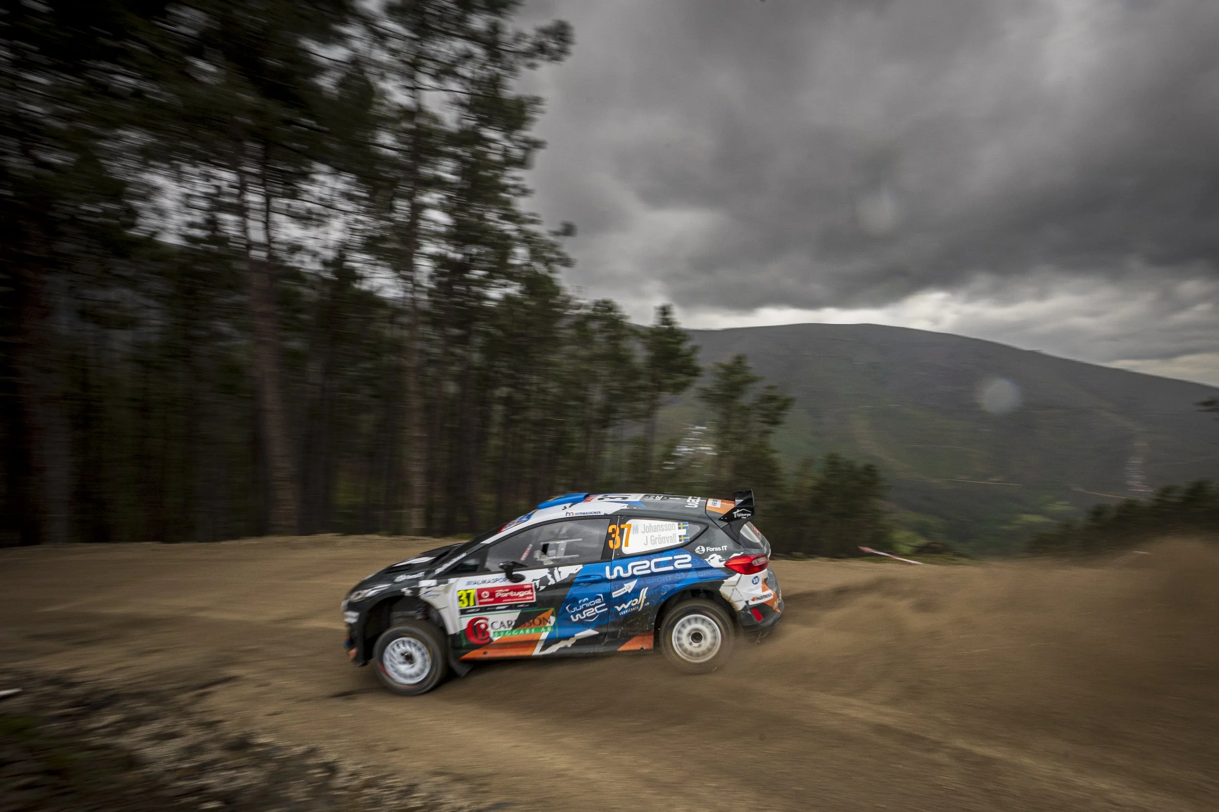A rally car with various sponsor decals racing on a dirt track with a mountainous landscape and dark, cloudy sky in the background.