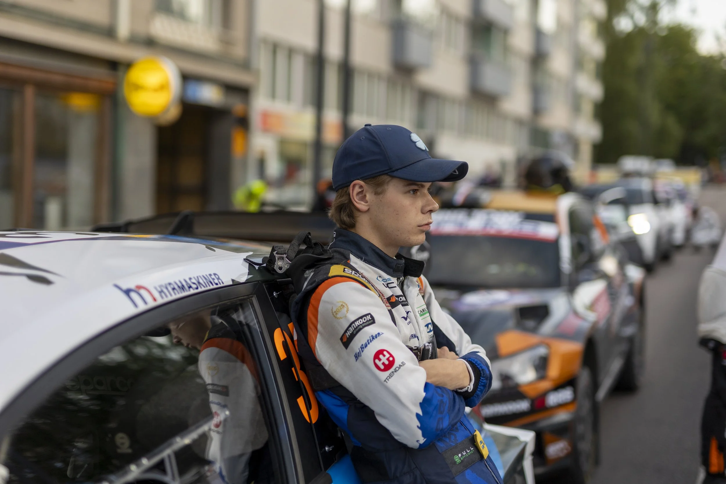 A young male race car driver with crossed arms stands beside his race car in a pit area, wearing a racing suit and cap, with other race cars and buildings in the background.