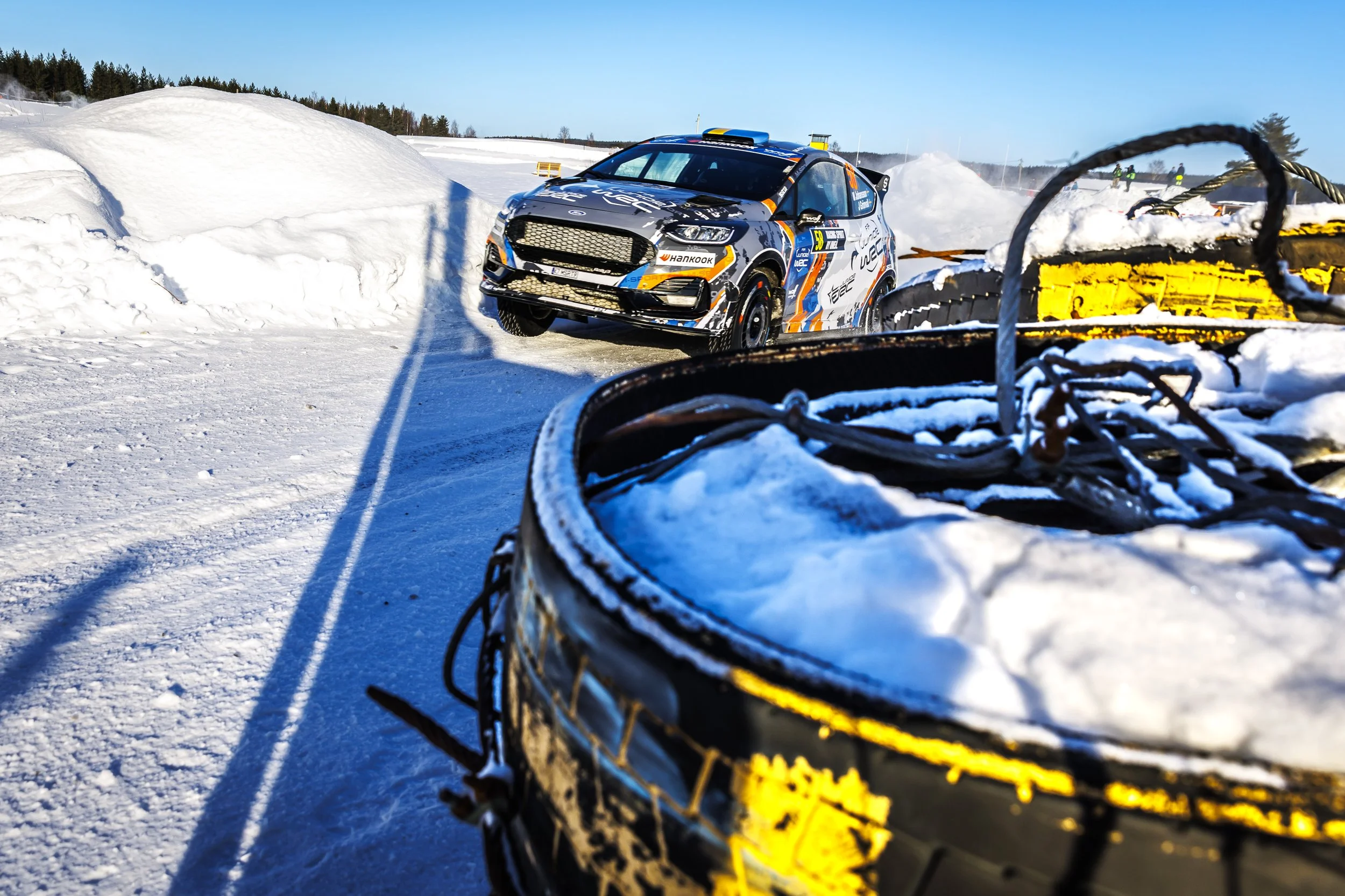 A rally car driving on snow with snowbanks and a clear blue sky in the background, seen from a low angle near snow-clearing equipment.