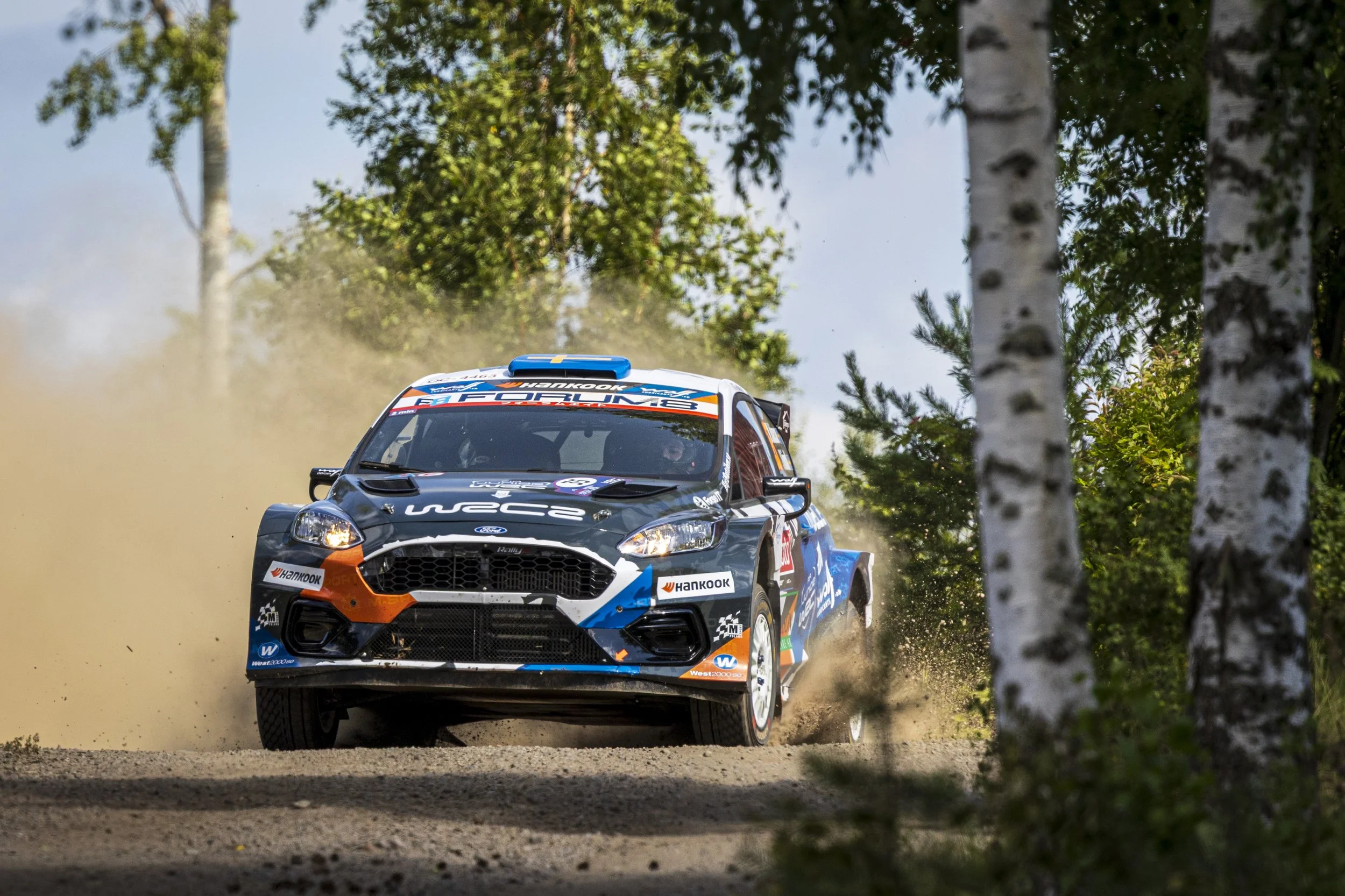 A rally car speeding on a dirt road through a wooded area, kicking up dust as it navigates a turn.