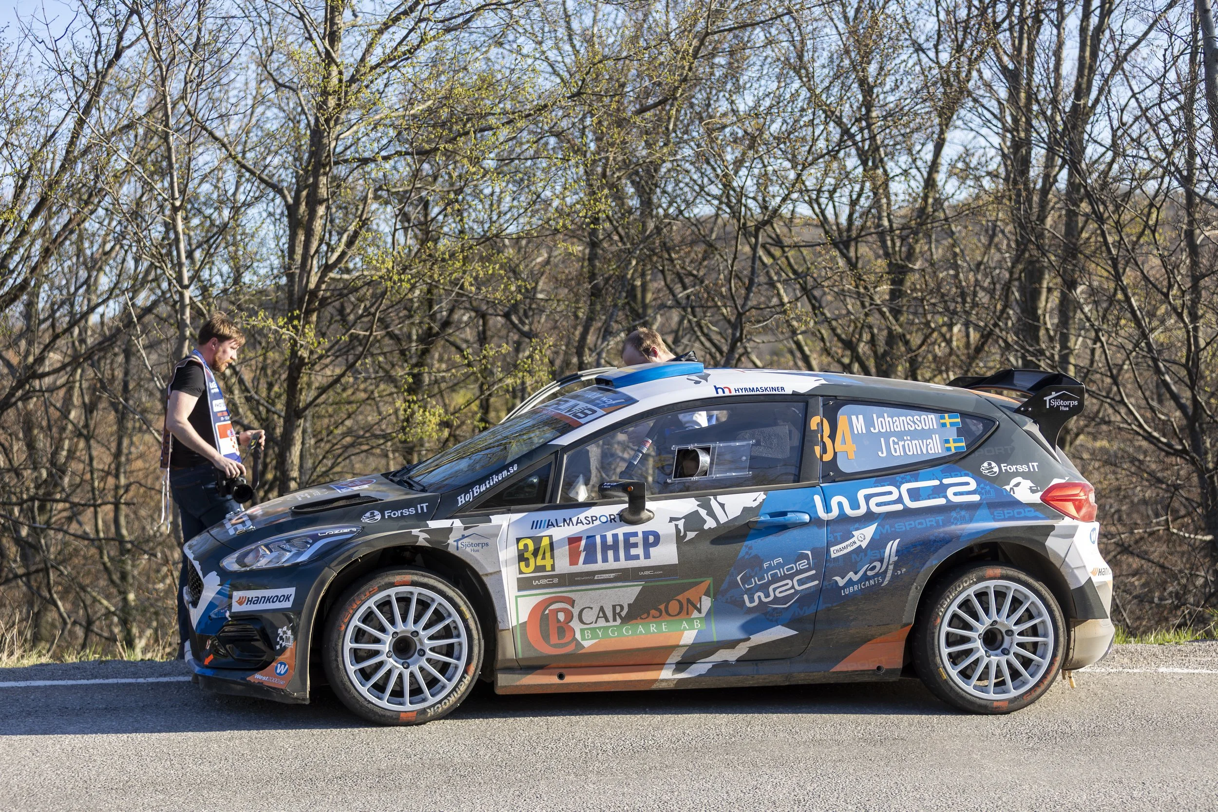 A rally car with racing decals and sponsorships parked on the side of a road, with two men inspecting or working on it, trees and a blue sky in the background.