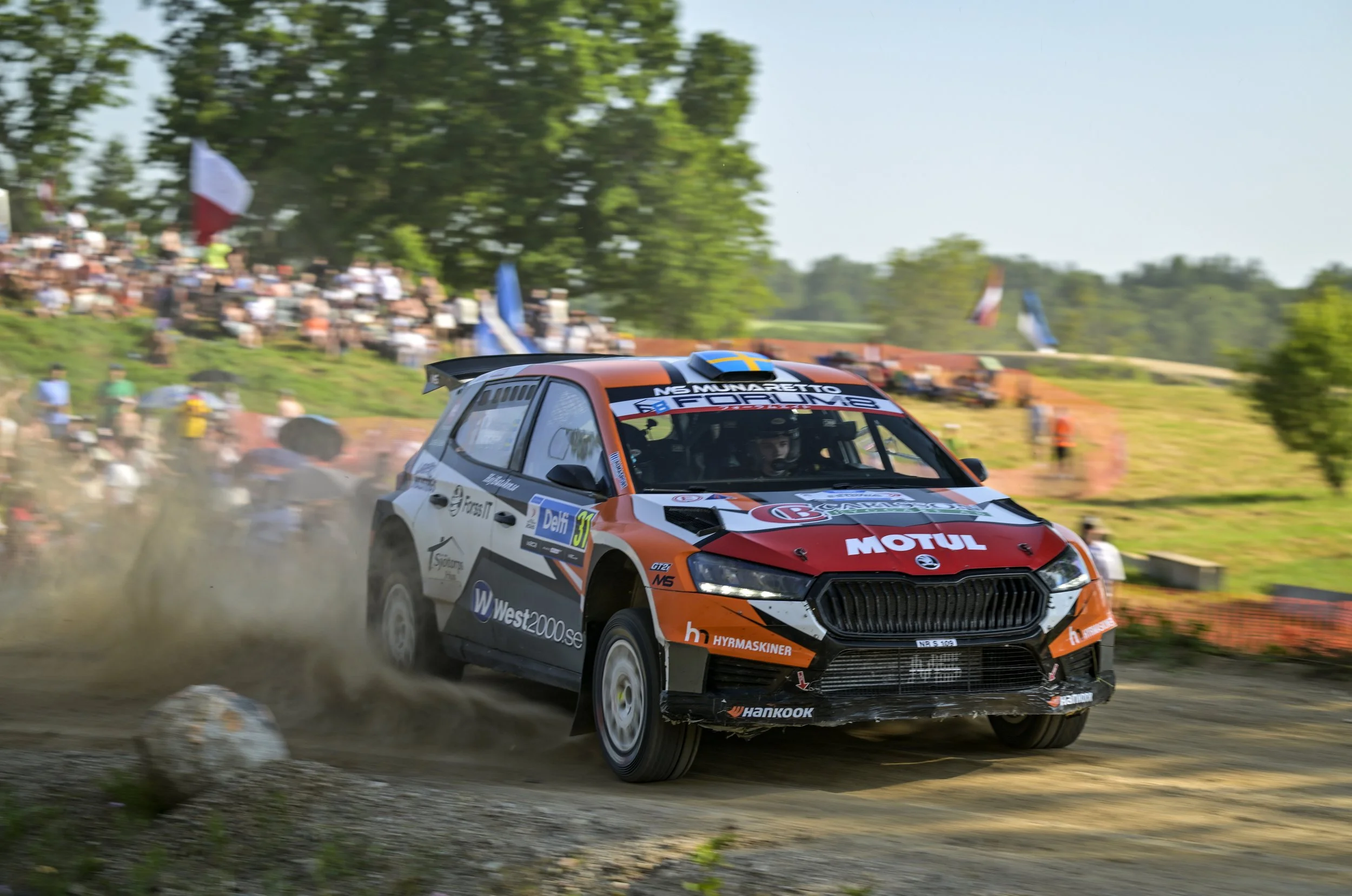 A rally car speeding on a dirt track, with a crowd of spectators in the background.