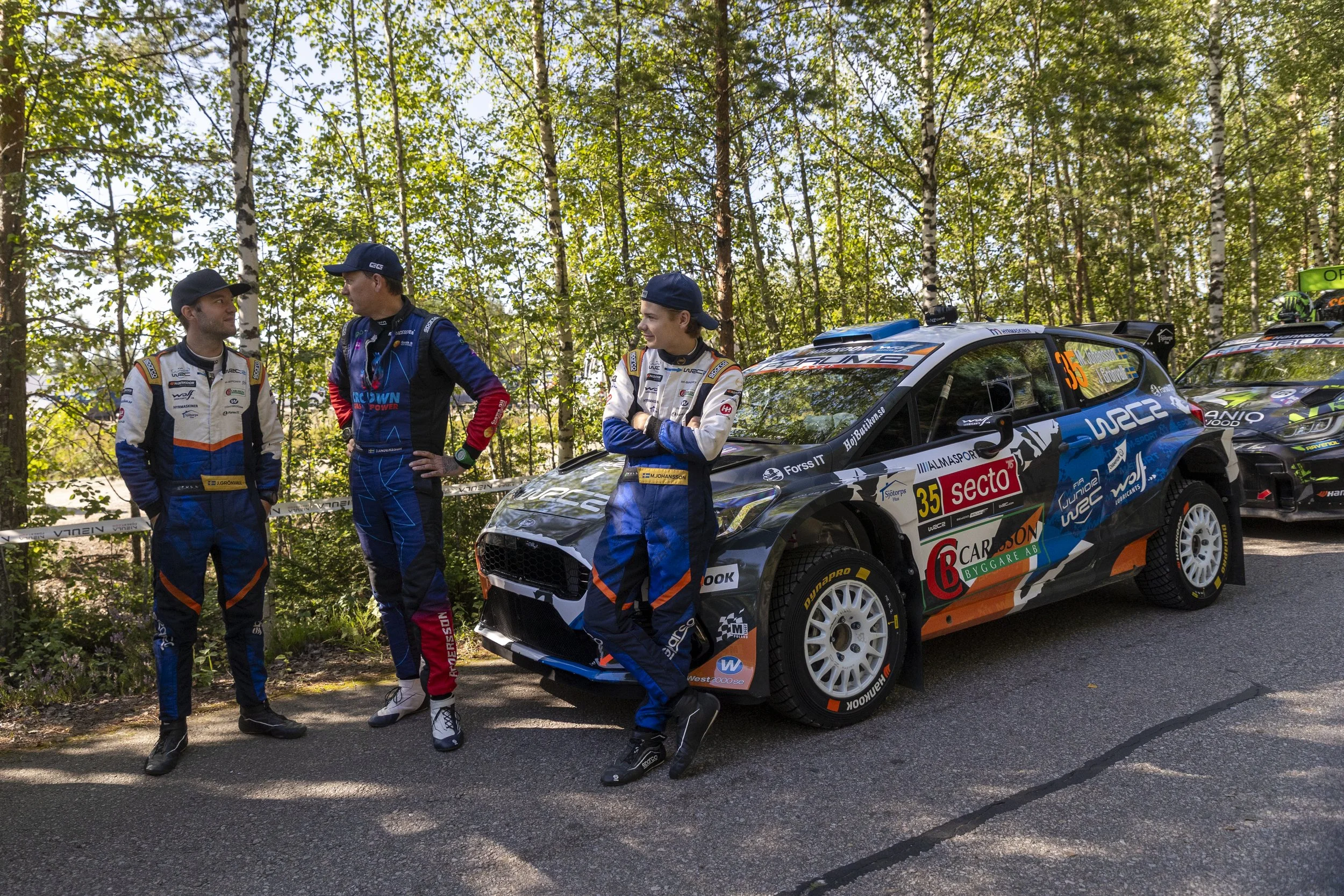 Three rally drivers in racing suits standing beside a rally car with sponsor decals, parked among trees during daytime.