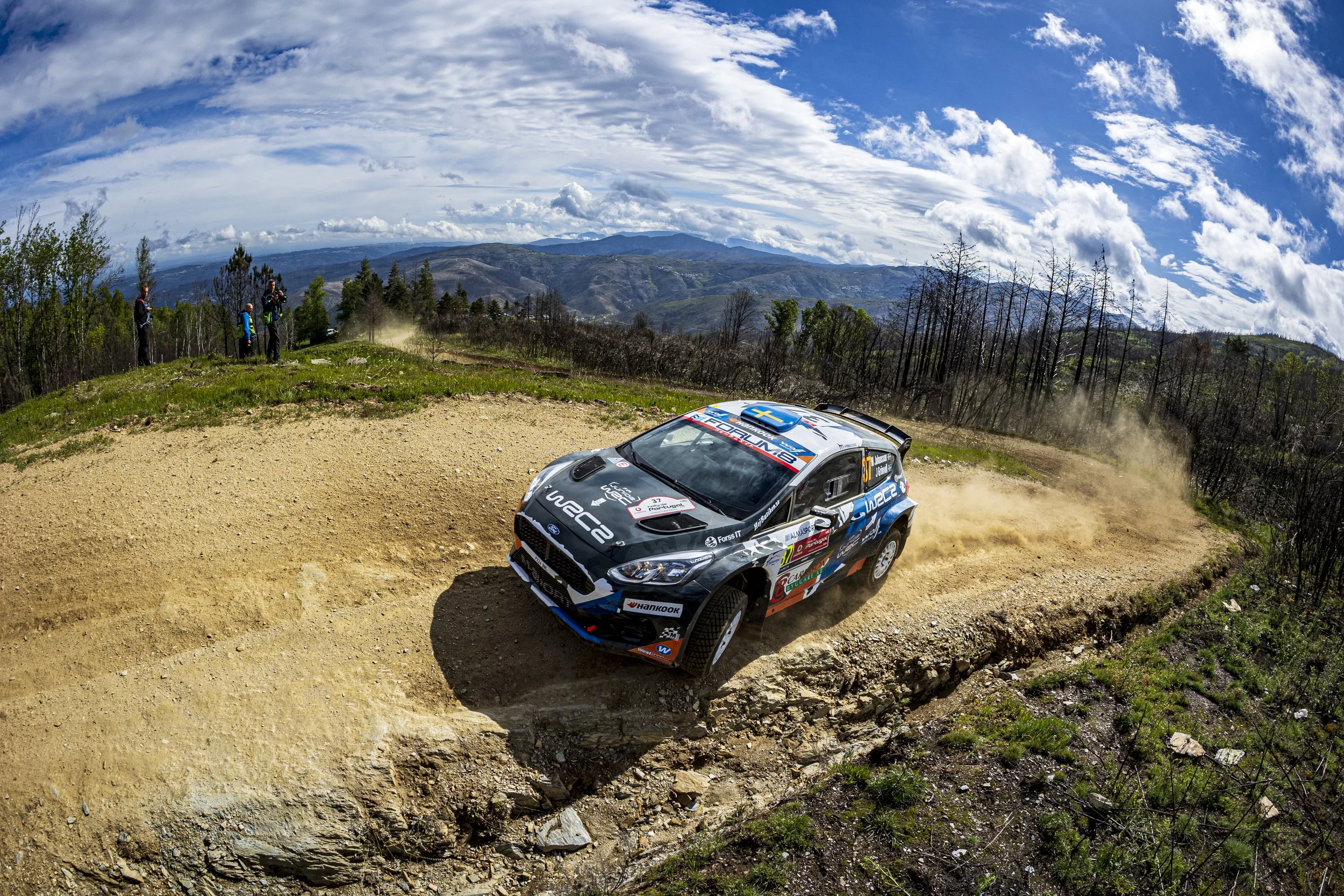 A rally car speeding on a dirt road in a mountainous landscape with a partly cloudy sky and a few spectators on the side.