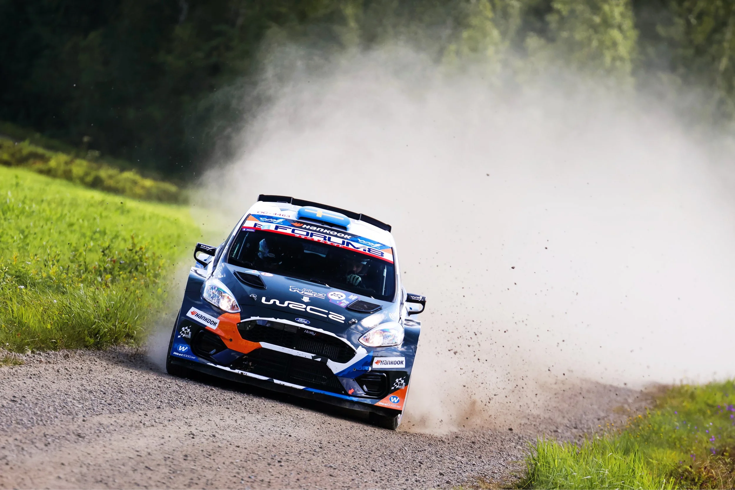 A rally car speeding along a gravel road, kicking up dust and dirt, with a lush green forest in the background.