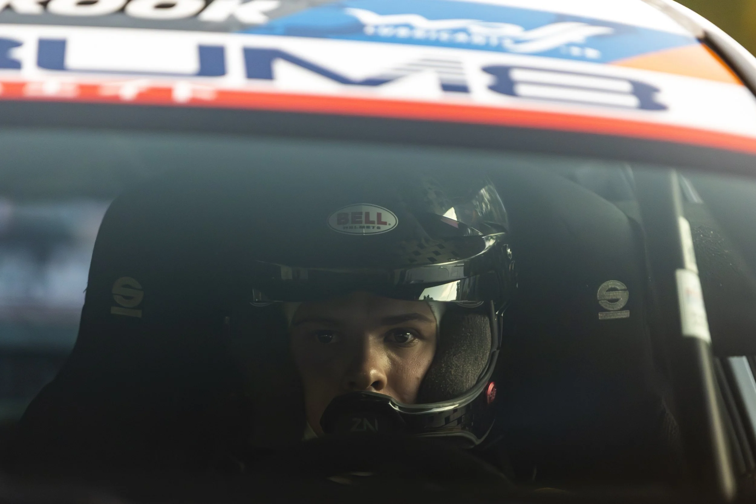 A close-up of a person's face reflected in a mirror while wearing a racing helmet, with the background showing part of a car with racing decals.