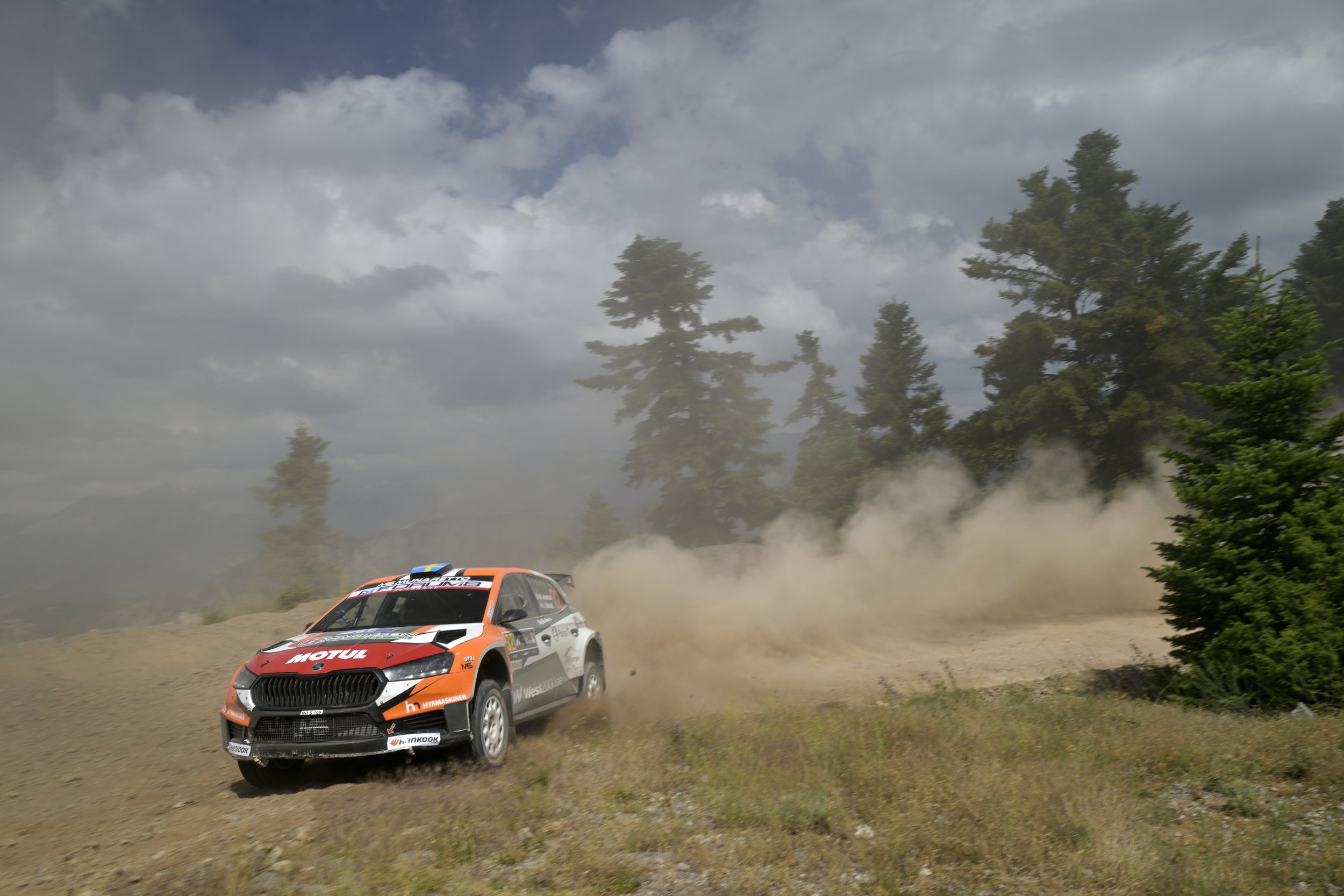A rally car racing on a dirt road through trees with a cloud of dust behind it.