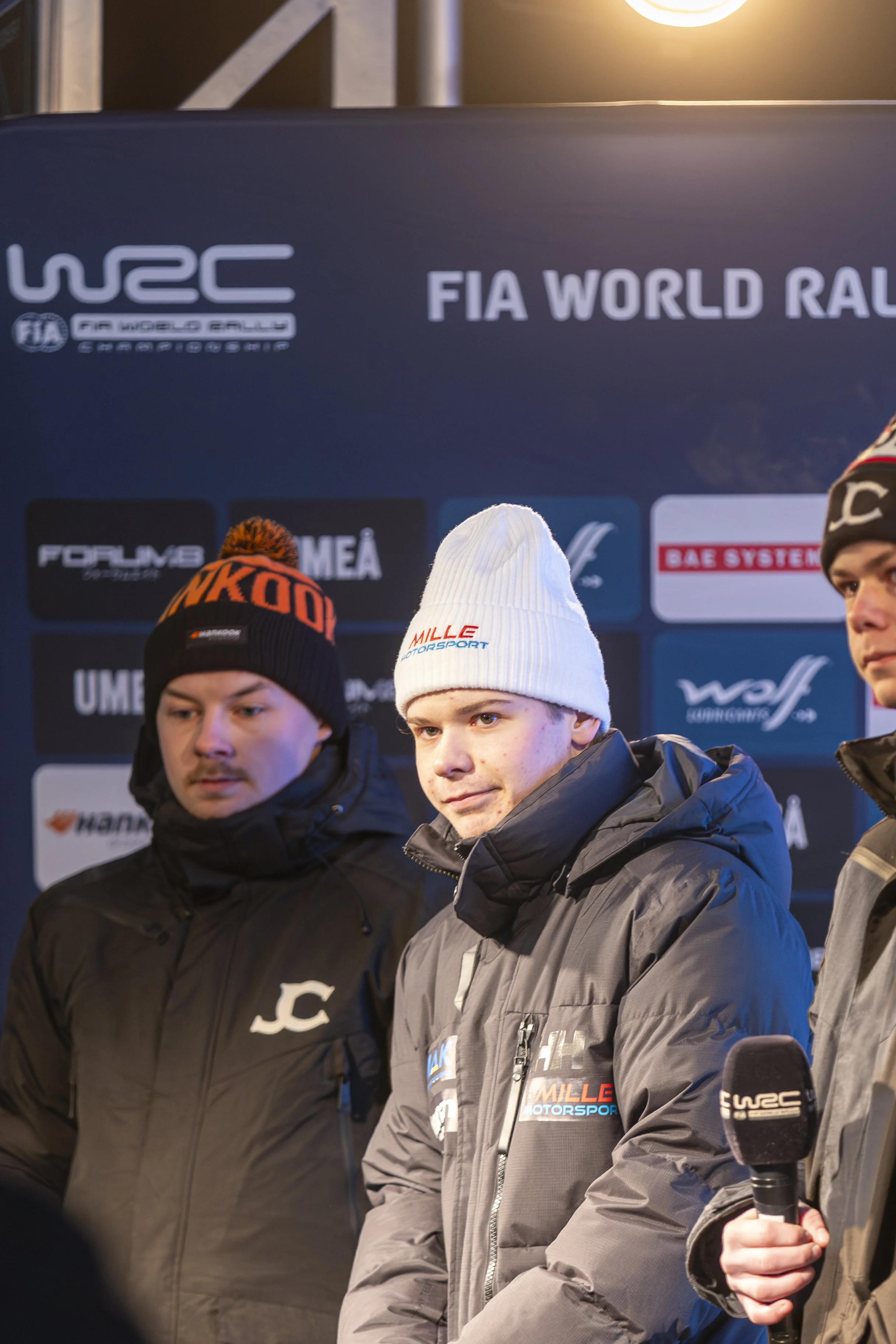 A group of young men in winter jackets and hats standing in front of a blue banner with logos and text including 'FIA WORLD RALLY' and 'WRC'.