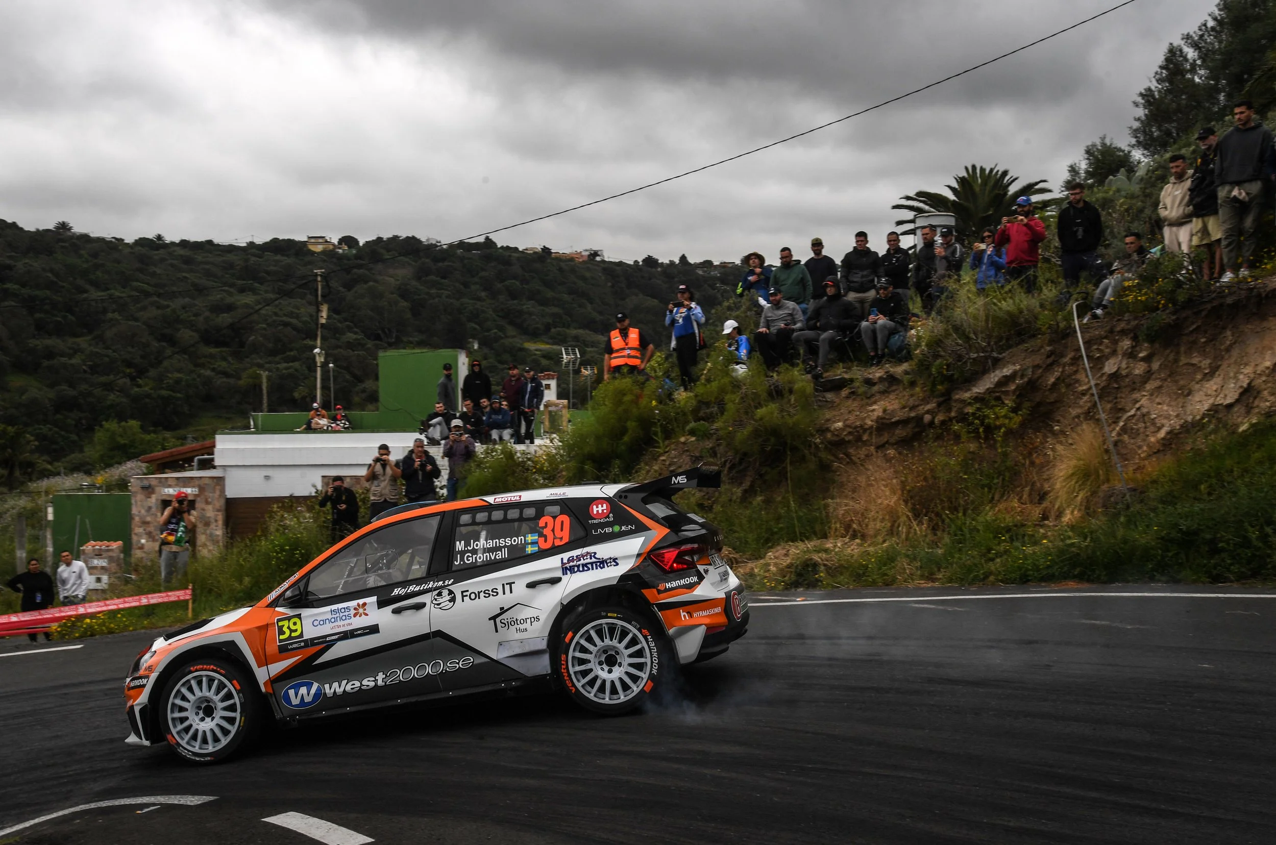 A rally car with the number 39 is racing on a winding road amidst a crowd of spectators watching from the side on a cloudy day.