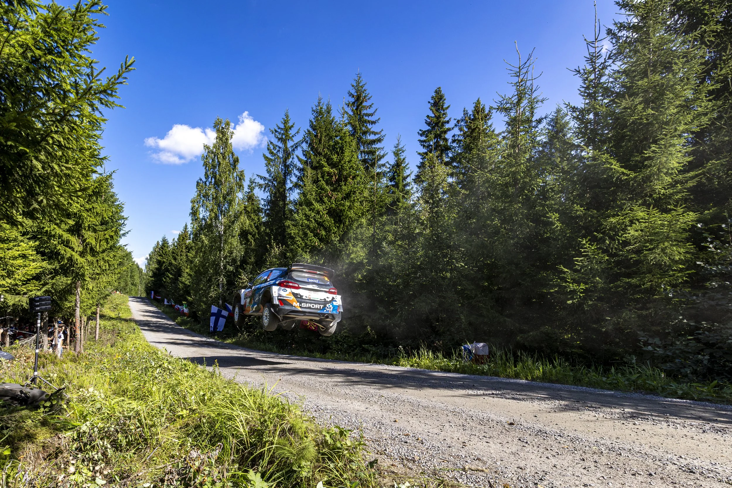 A rally car mid-air jumping over a dirt road surrounded by green trees on a sunny day.