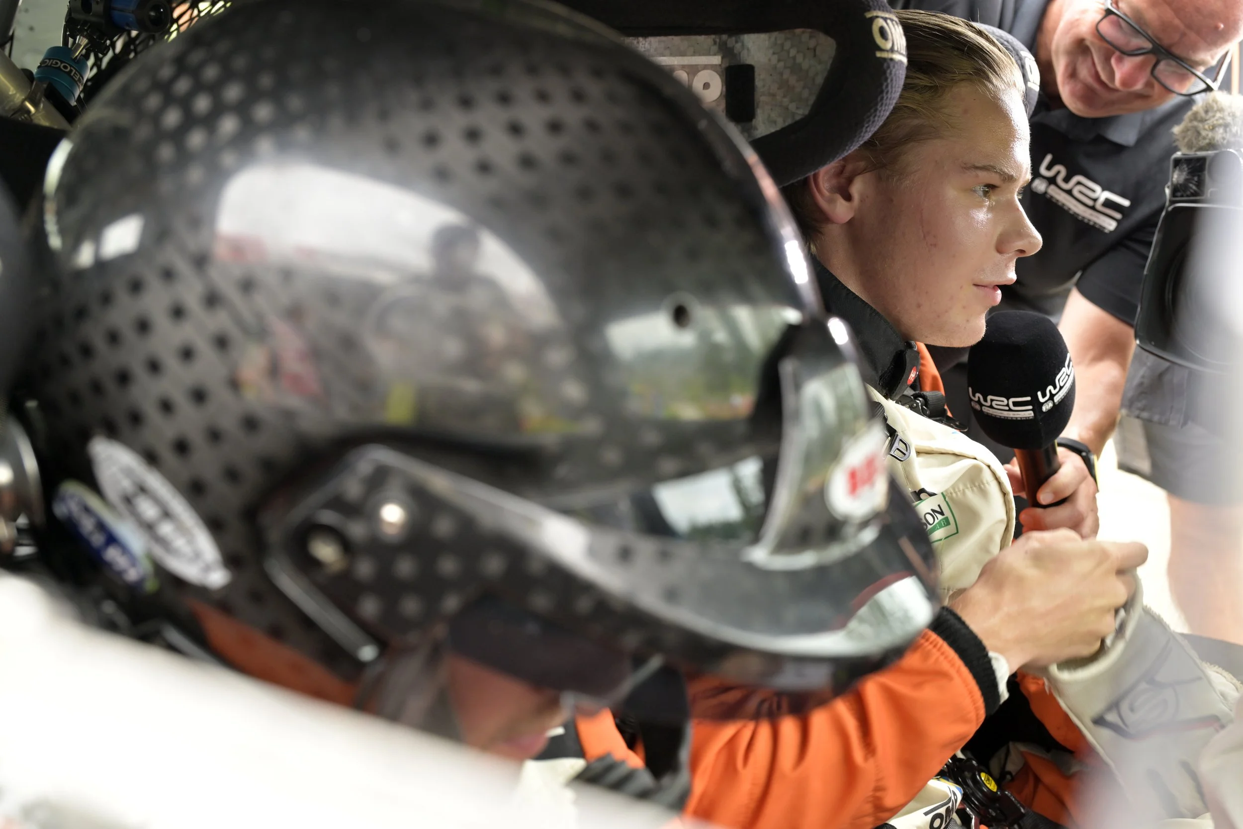 Close-up view of a racing helmet with a helmet visor reflecting the scene, and a young male rally driver inside the car with a concerned expression, holding a microphone, while an older man speaks to him, standing outside the vehicle.