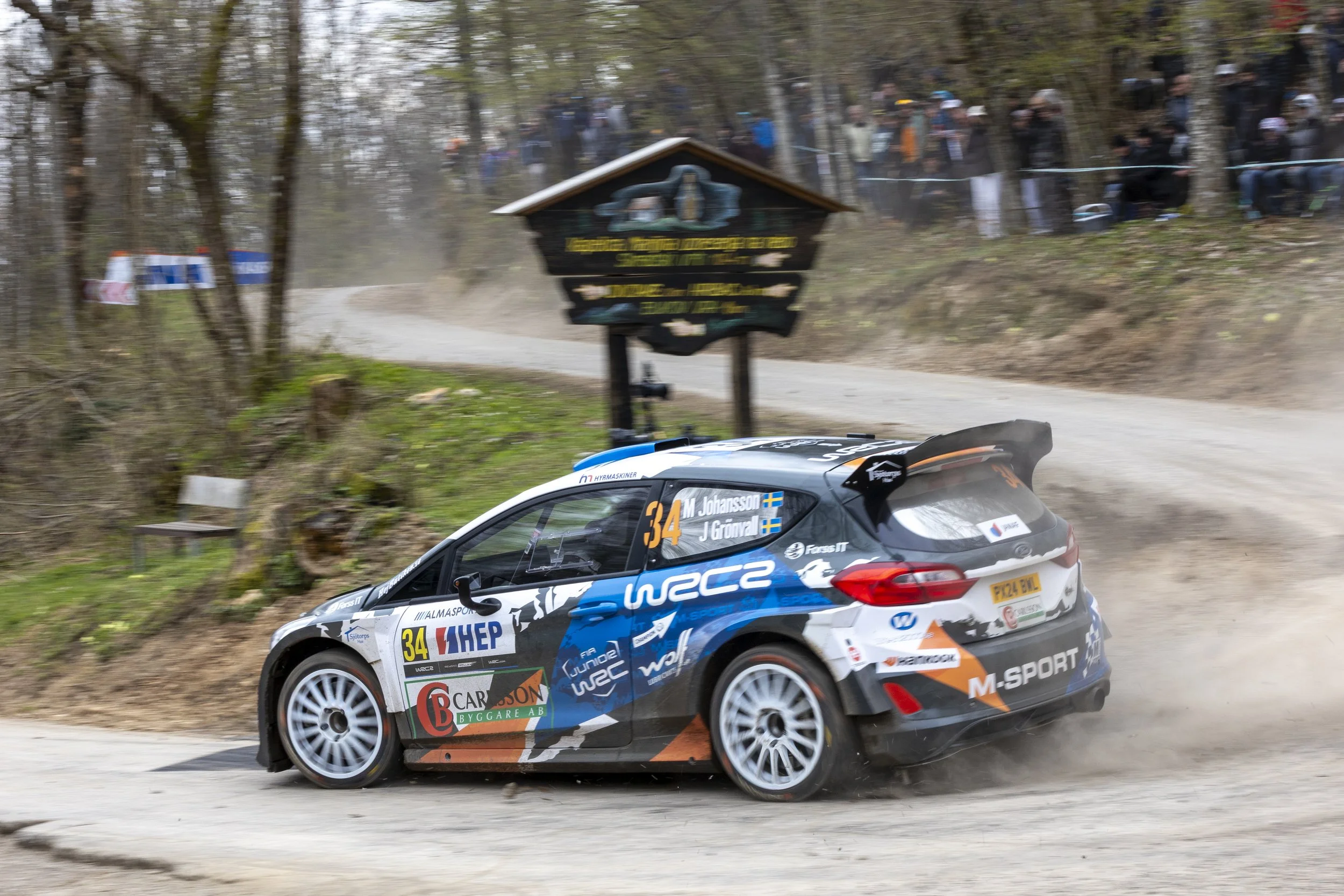 A rally car speeding on a dirt road with dust trailing behind, surrounded by trees and a crowd of spectators watching from the side of the trail.