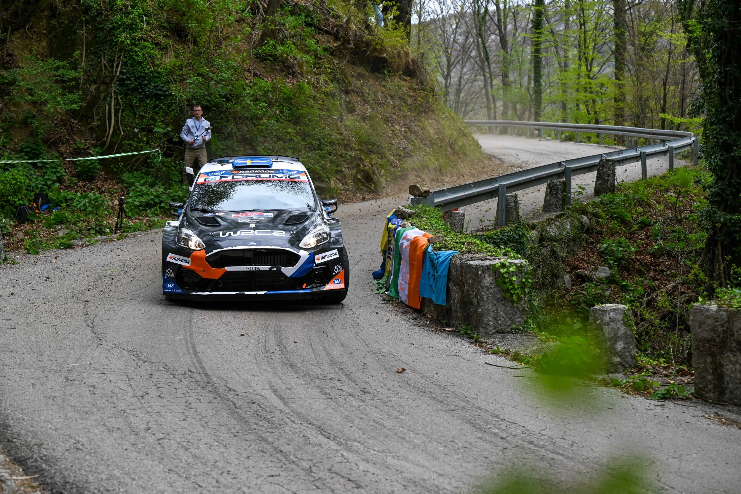 A rally car parked on a narrow mountain road with a roadside barrier and colorful towels hanging on the guardrail. A person stands nearby, holding a clipboard, in a wooded area with trees and green foliage.