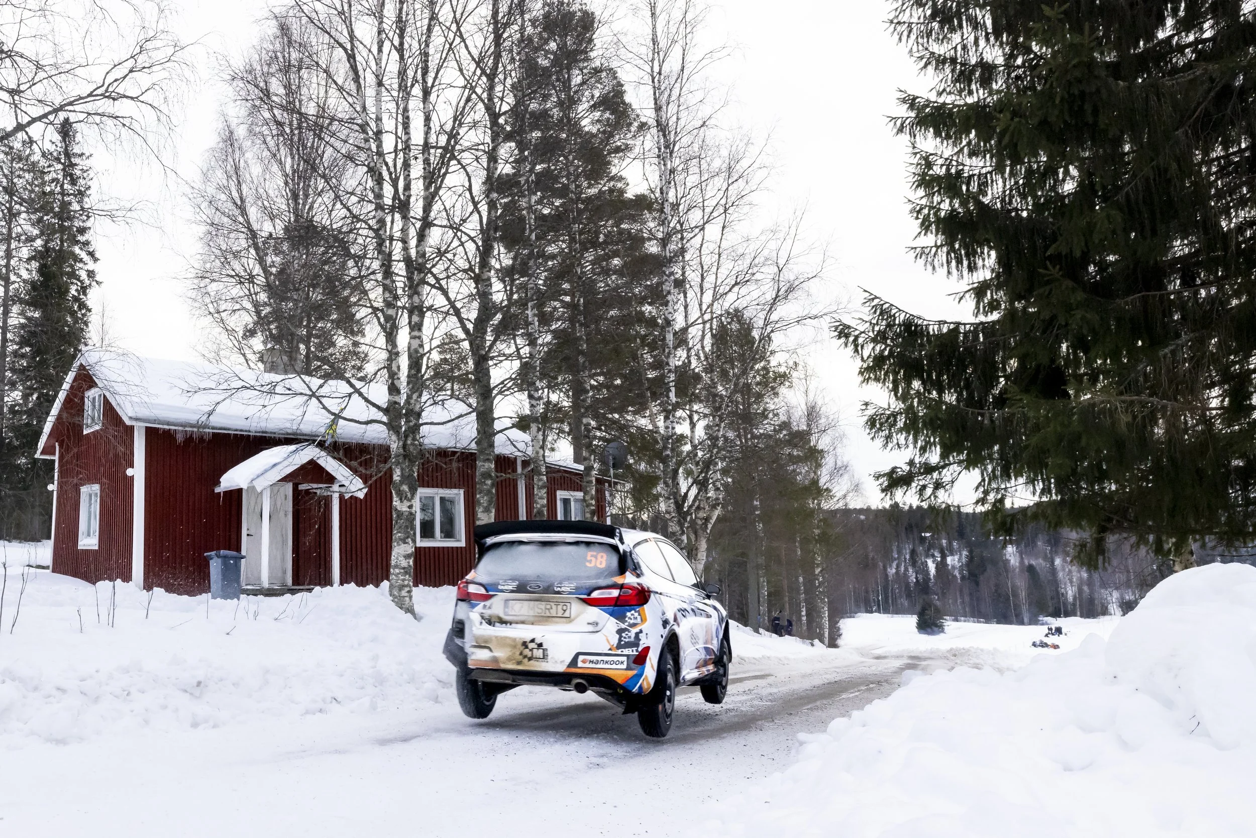 A snowy landscape with a red house, leafless trees, and a car parked on the snow-covered road.
