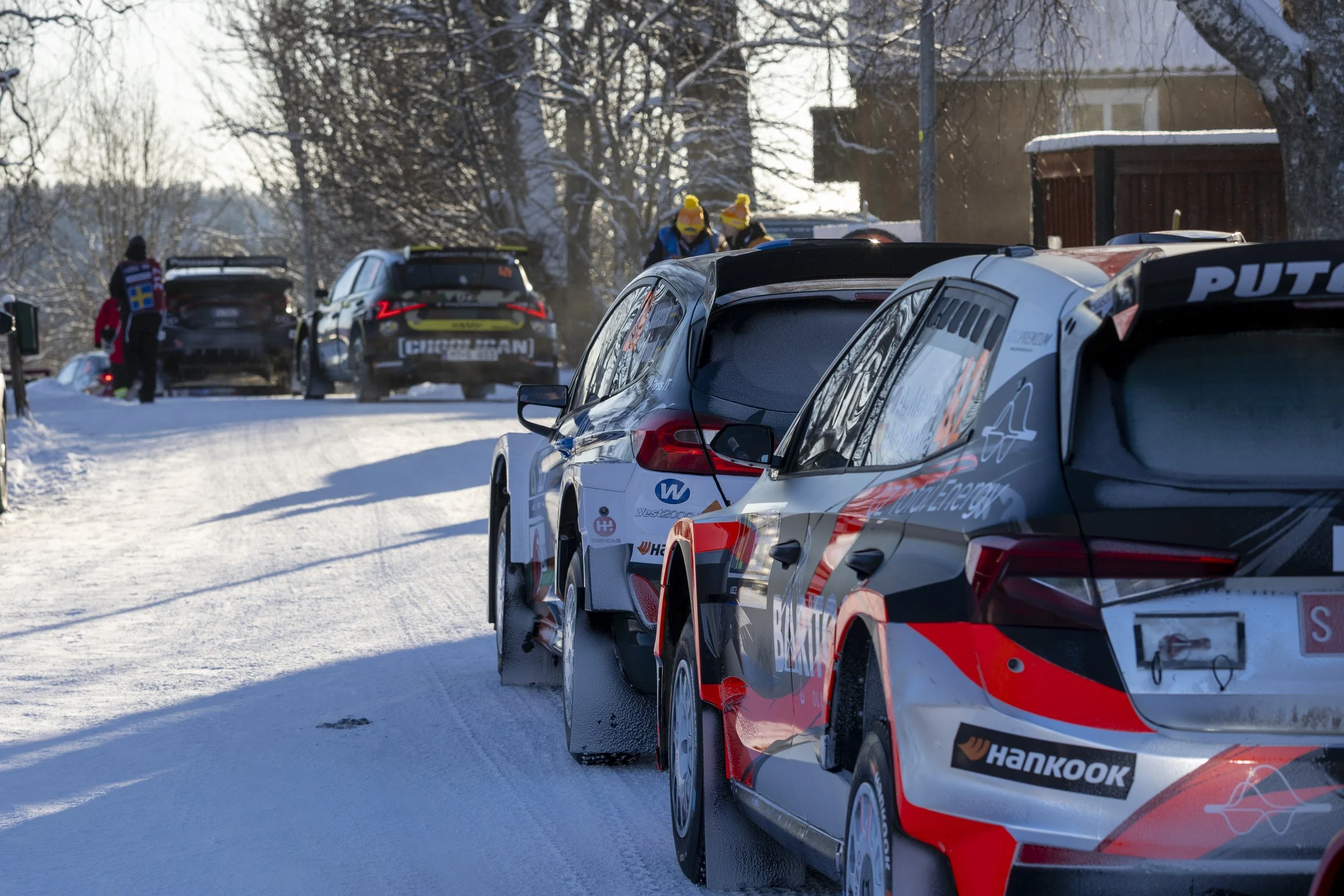 Race cars parked on a snowy road with people in winter clothing and trees in the background.