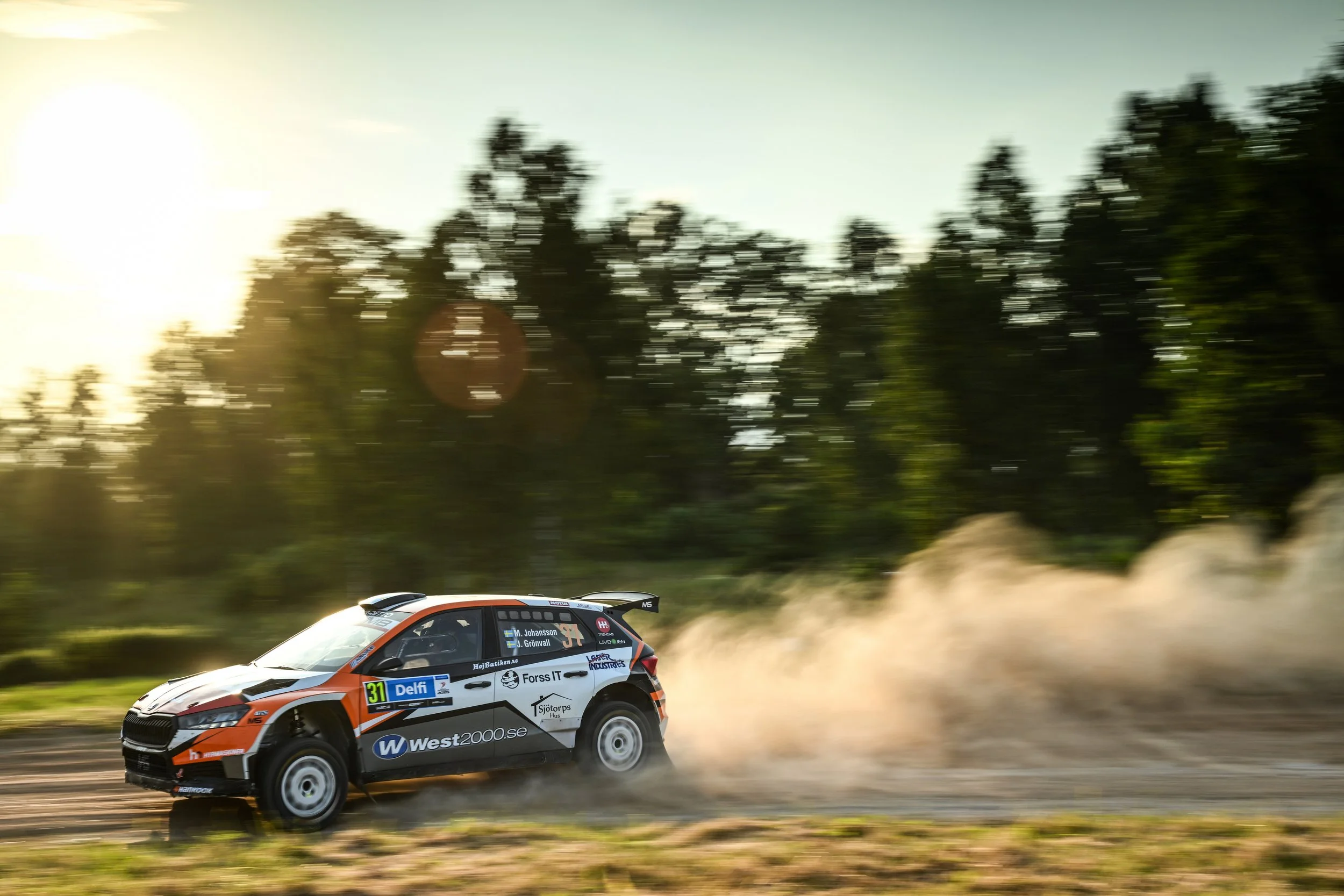 A rally car racing on a dirt road, kicking up dust, with a forested background and the sunlight shining through the trees.