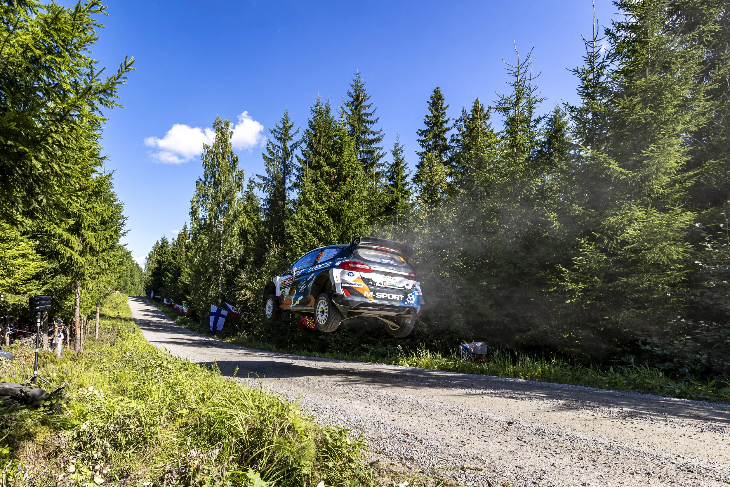 A rally car is airborne above a dirt road surrounded by dense green forest on a bright, sunny day.