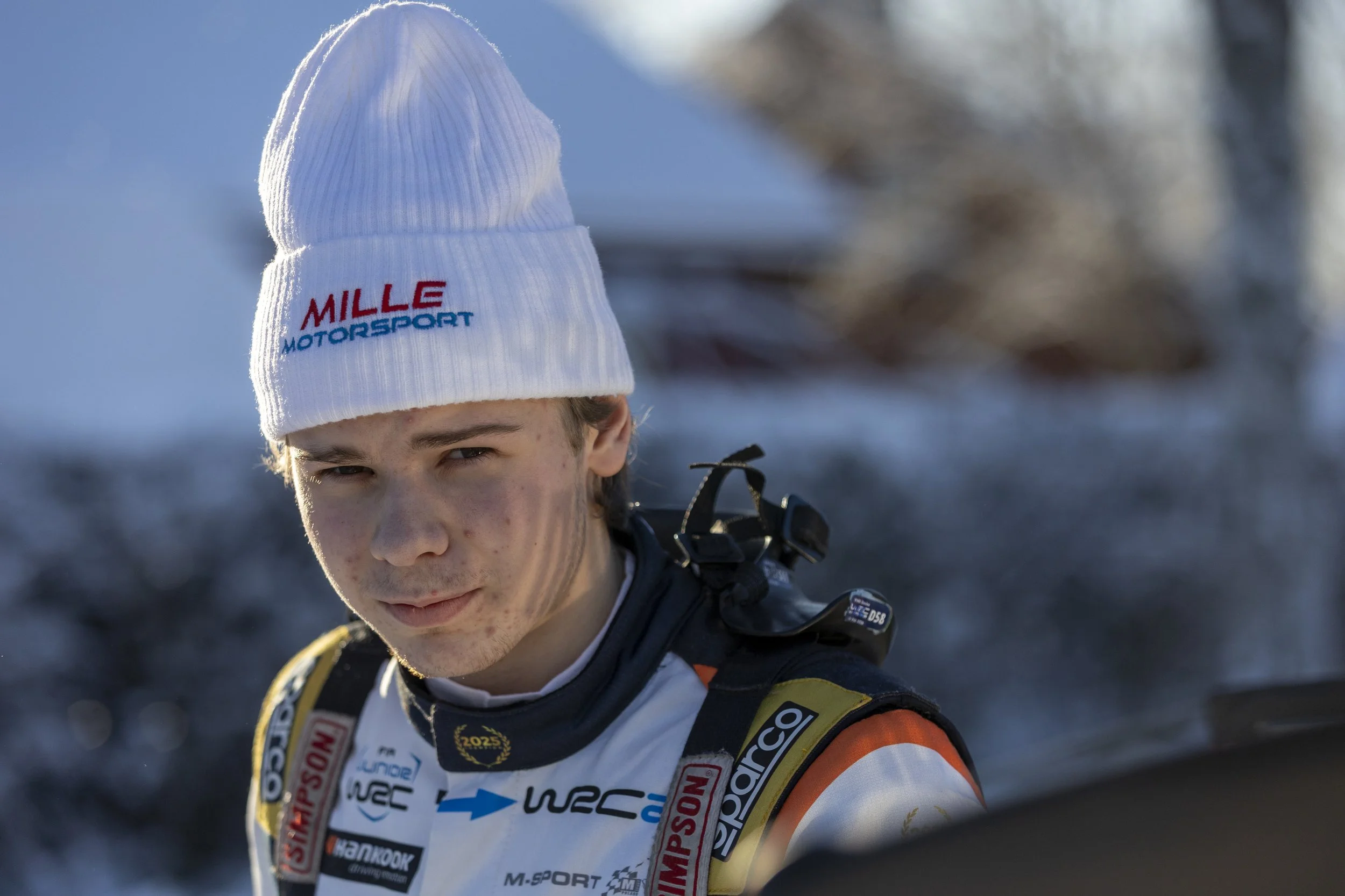 A young man wearing a white beanie with 'MILLE MOTORSPORT' embroidered in red and blue, dressed in a racing suit with various sponsor patches, outdoors in a snowy environment.