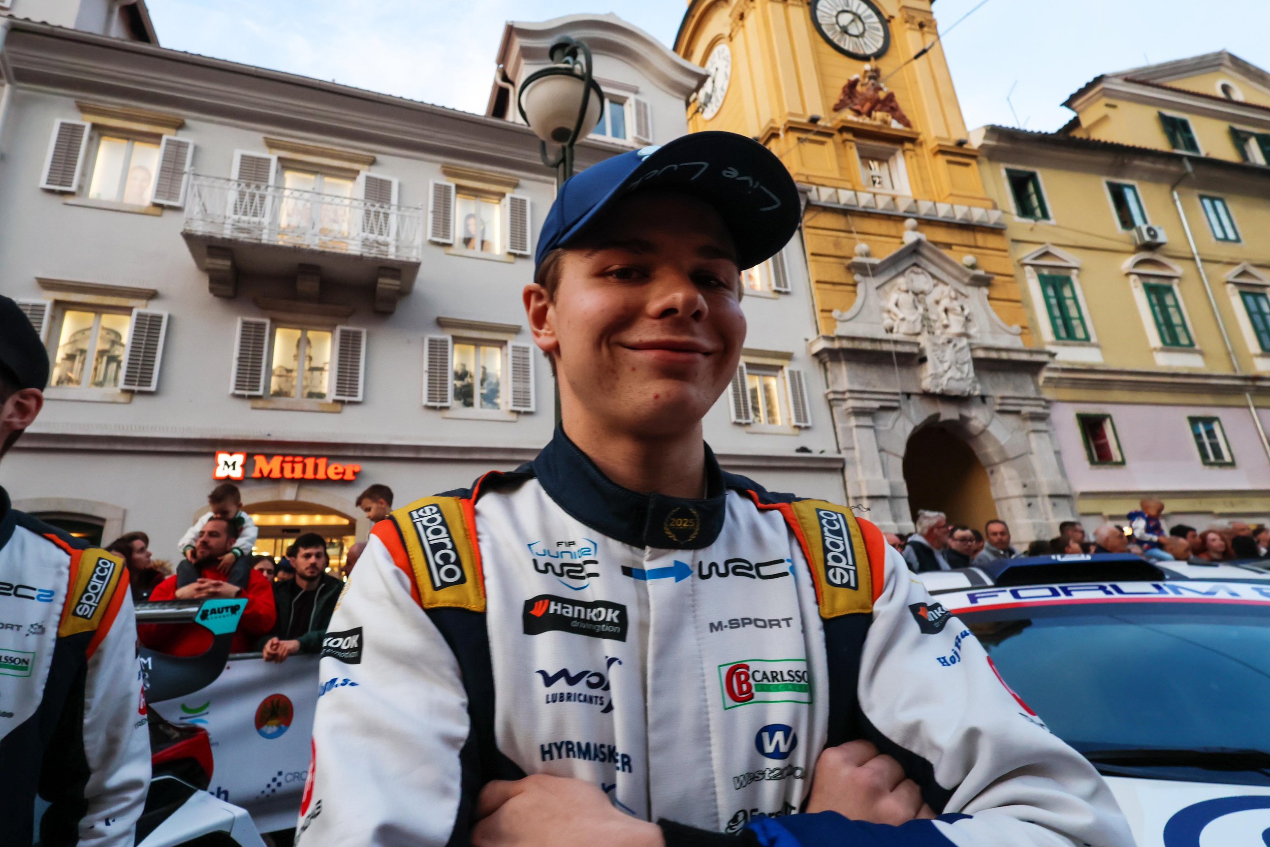 A young male race car driver taking a selfie in front of a historic European town square with colorful buildings and a clock tower.