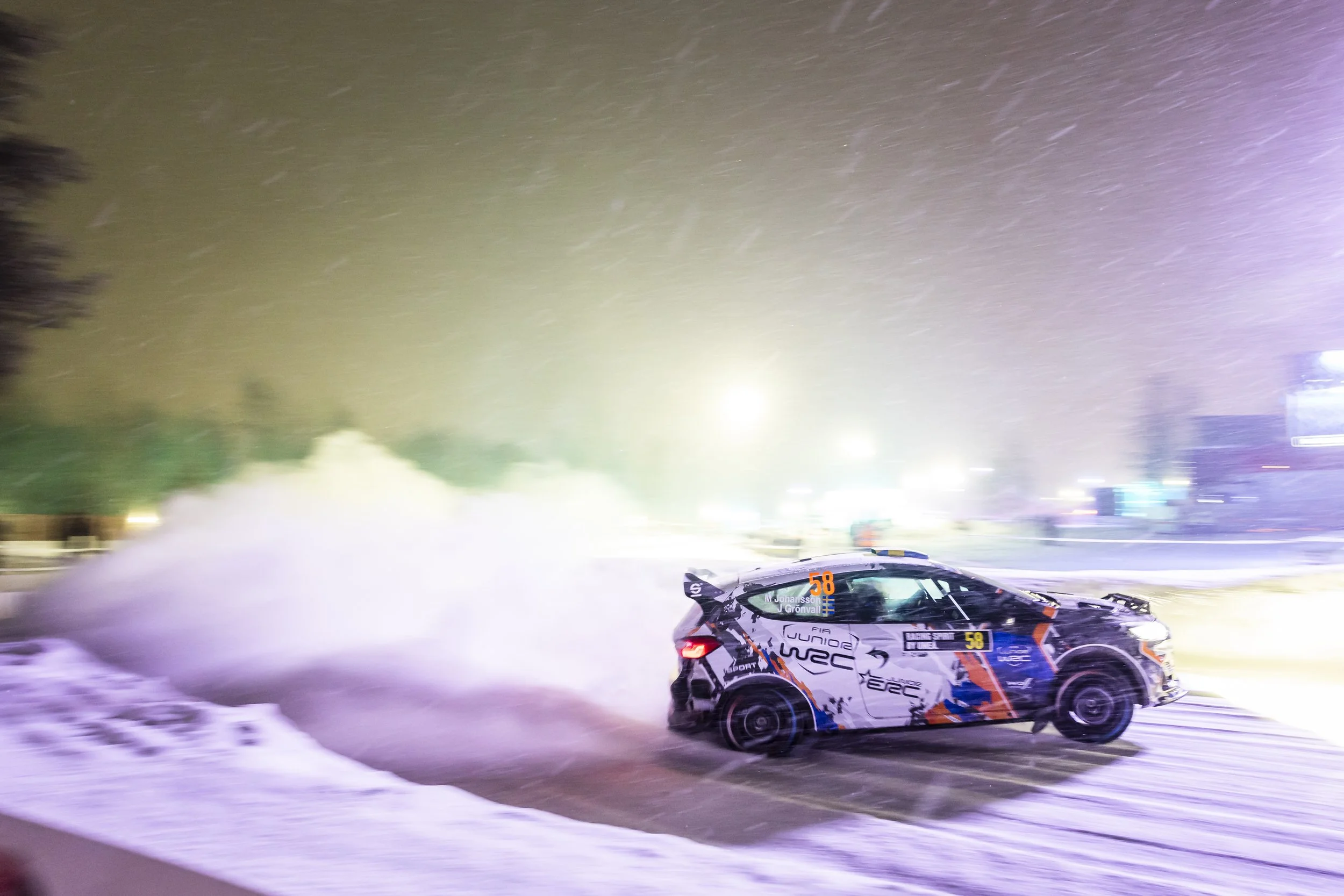 A rally car racing on a snowy track at night, with snow being kicked up behind it and bright lights illuminating the scene, and a mountain or hill in the background.