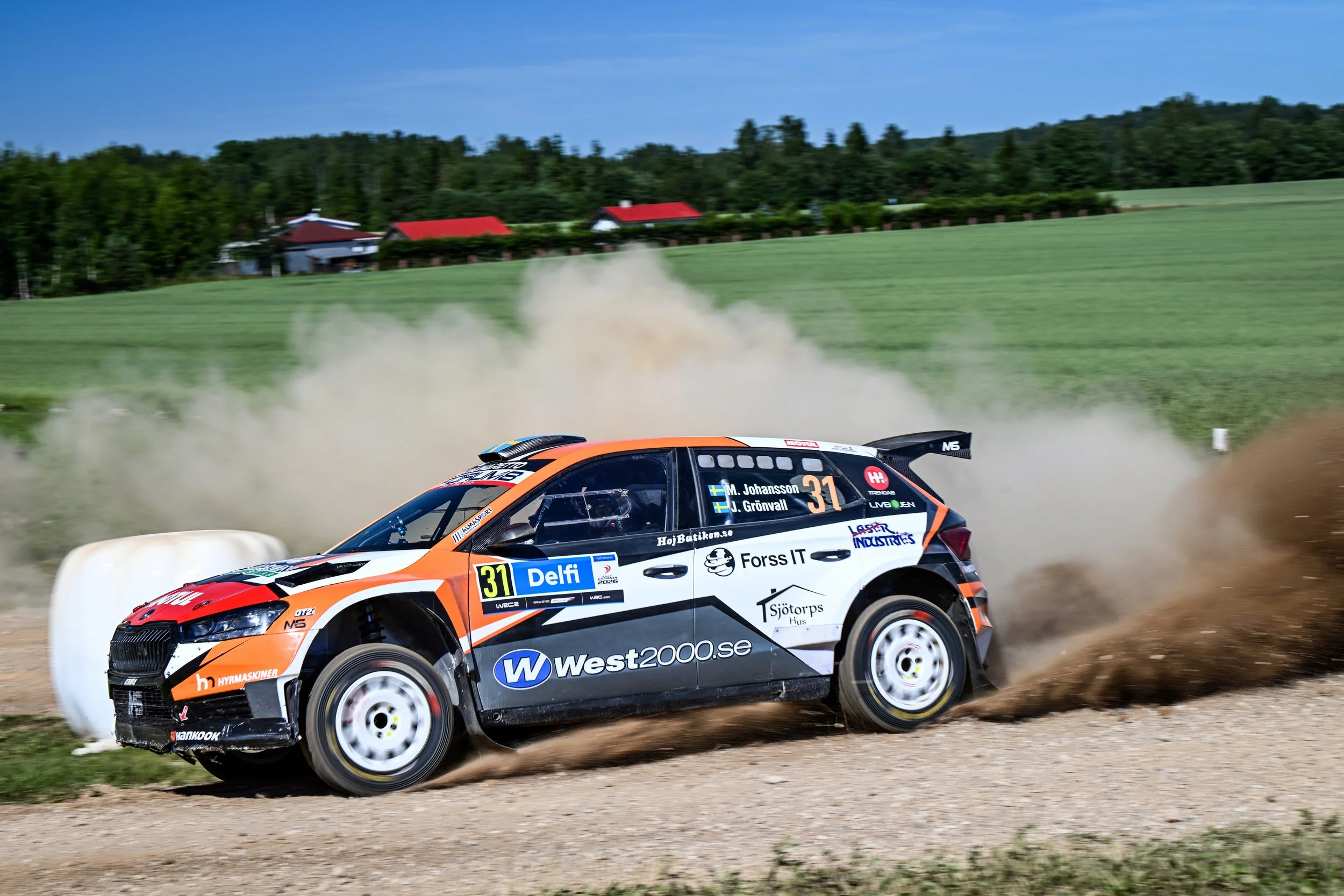A rally car speeding on a dirt track kicking up dust, with green fields and trees in the background under a blue sky.