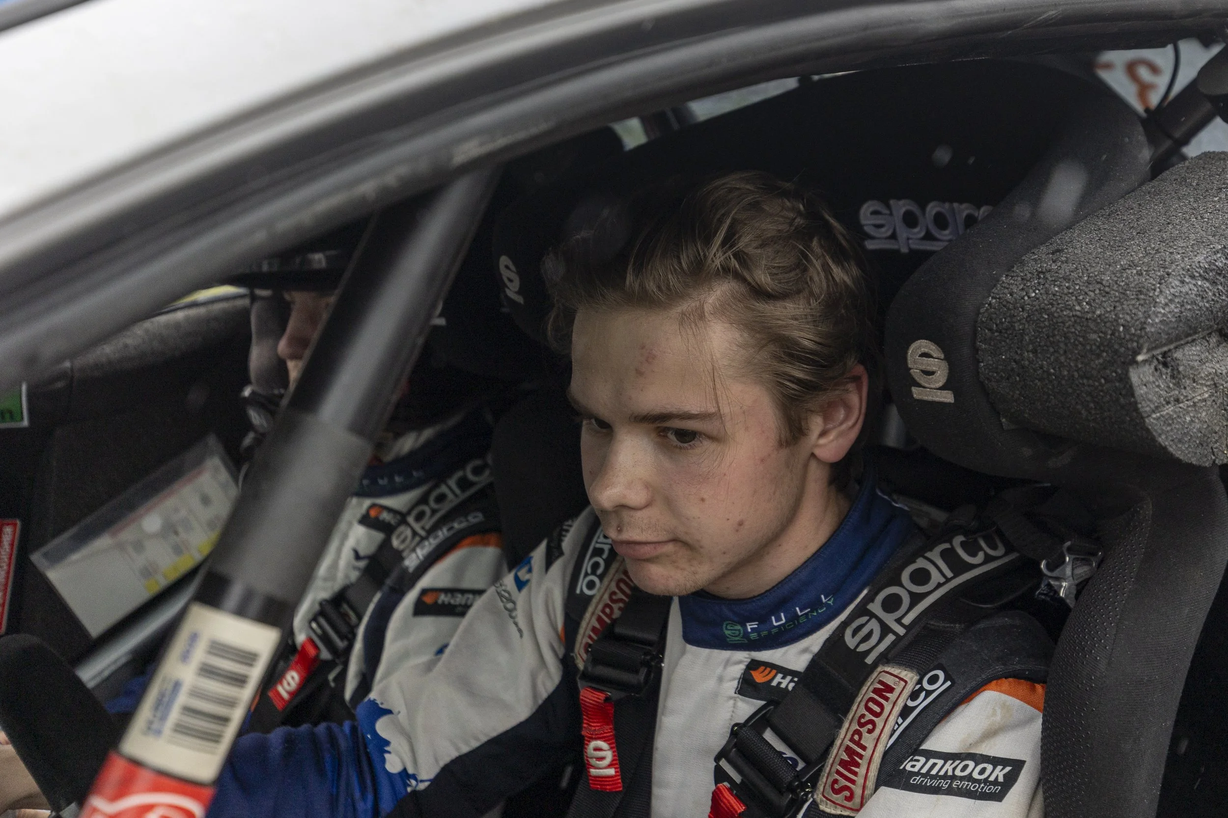 Close-up of a young race car driver with light brown hair, sitting inside a race car with a helmet and racing suit.