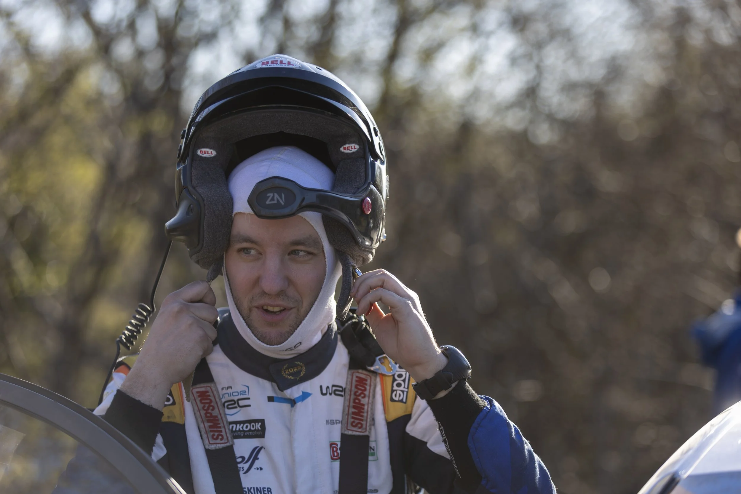 Man wearing a motocross helmet and racing suit adjusting his helmet strap outdoors with blurred trees in the background.