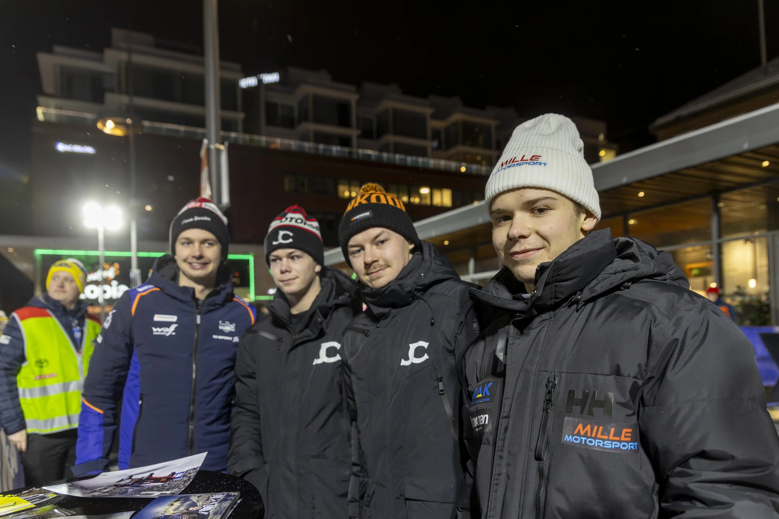 Group of young male race car drivers standing outdoors at night, wearing racing jackets and beanies, with a table of racing memorabilia in front of them and modern building in the background.