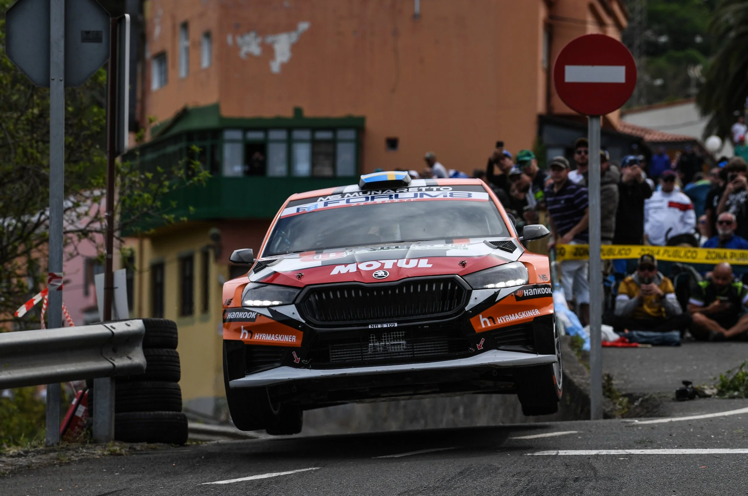A rally car in mid-air during a race, approaching a sharp turn with spectators watching from the side of the road. The car has sponsor logos, including 'Motul' and 'Hankook', and is surrounded by safety barriers and signage.