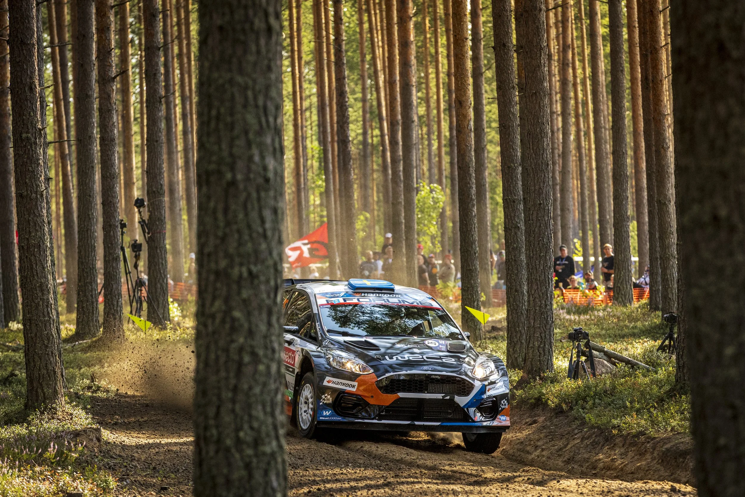 A rally car racing through a forest trail during a race, surrounded by tall trees and spectators watching from the sidelines.