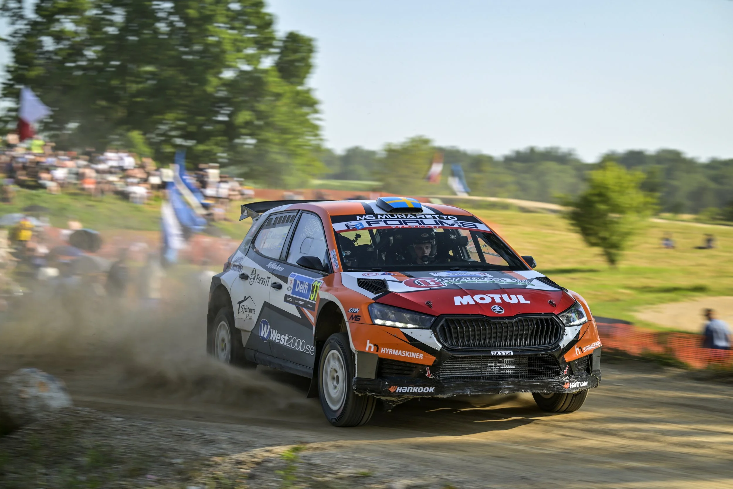 A rally car speeding on a dirt track, kicking up dust, with spectators and flags in the background.