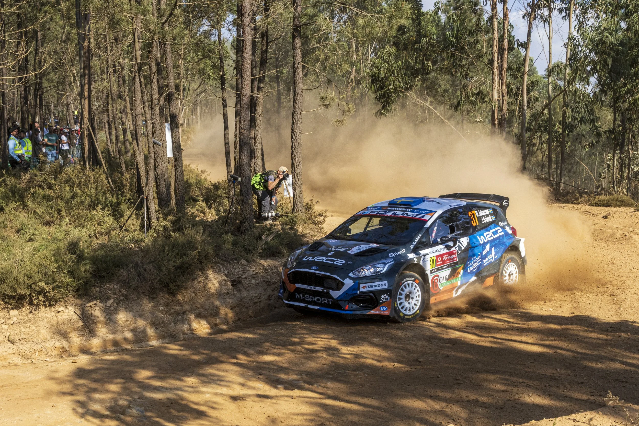 A rally car racing on a dusty dirt road through a forest, with a crowd of spectators watching from the side and a photographer capturing the moment.