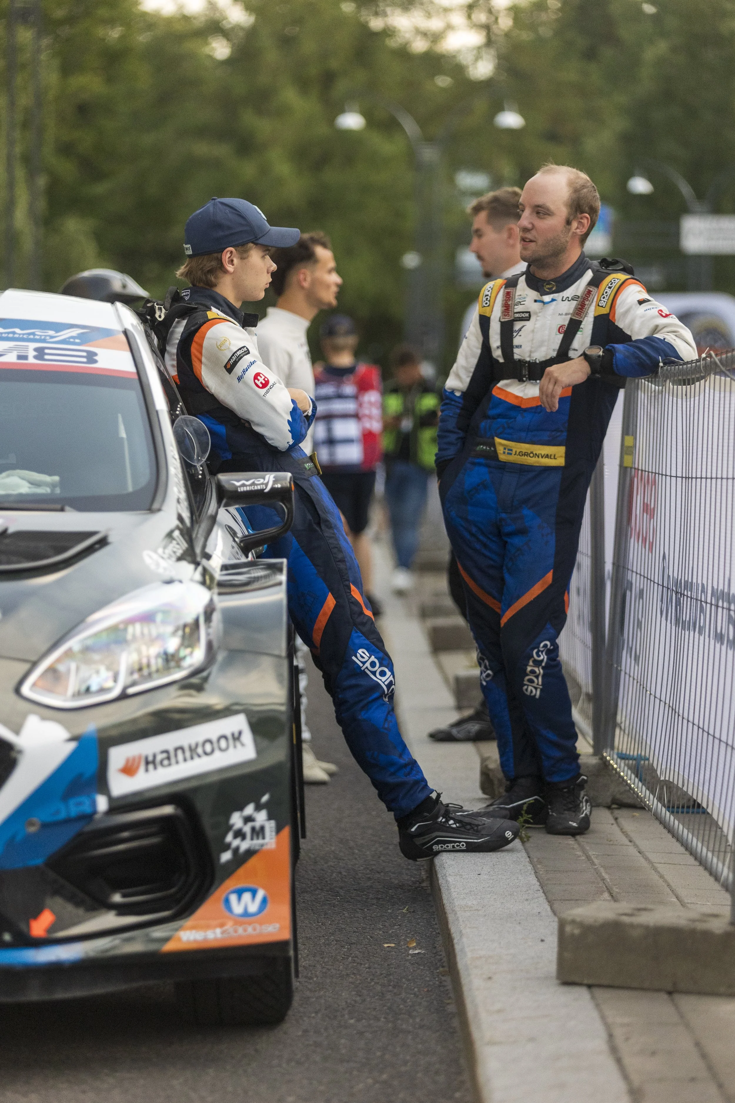 Two race car drivers in racing suits are standing and talking near a race car and a metal barricade, with spectators and trees in the background.