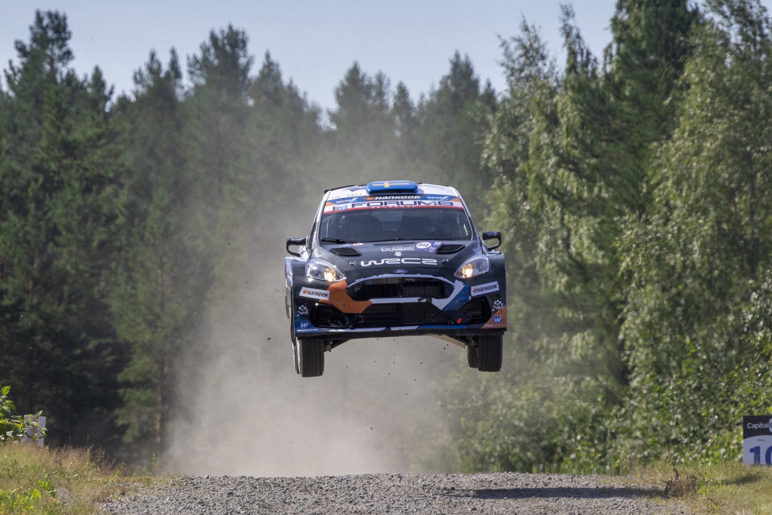 A rally car flying through the air on a dirt road surrounded by green trees.