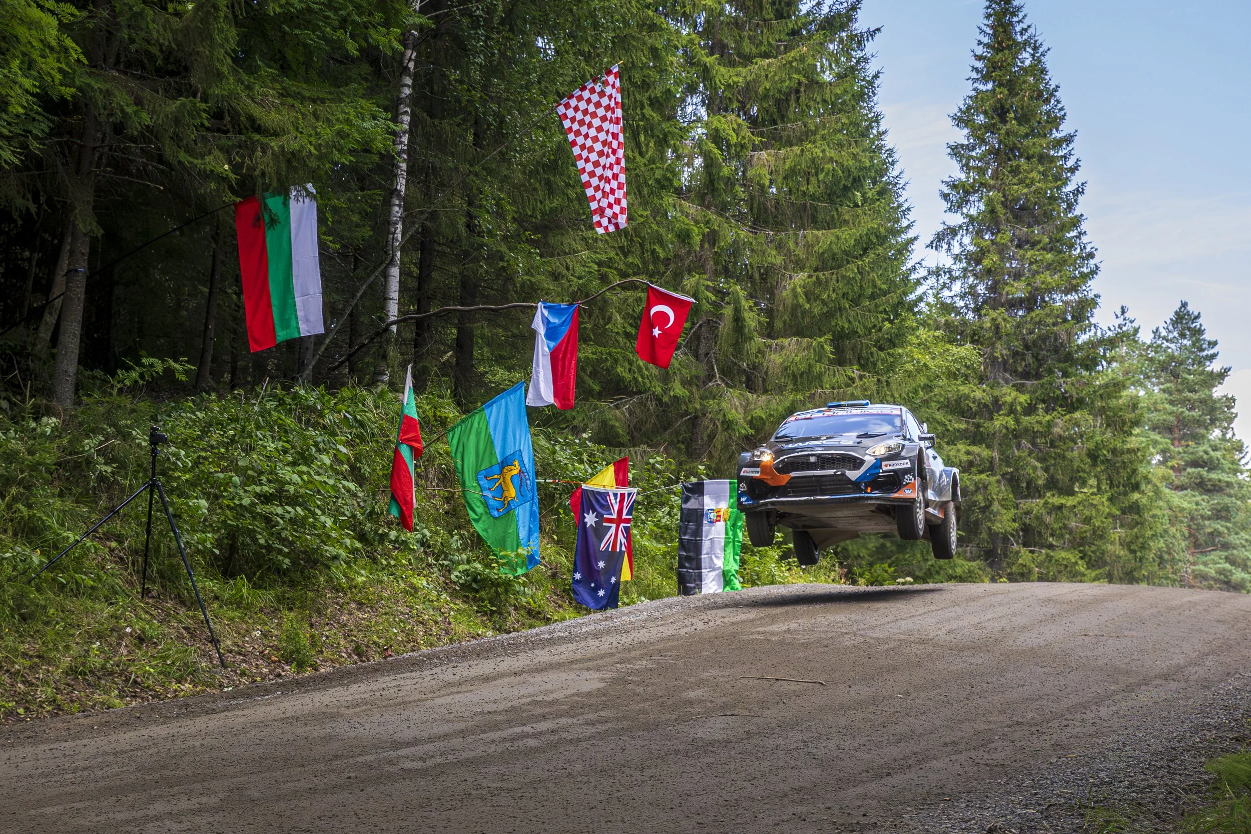 A rally car airborne over a dirt road with trees and multiple international flags hanging above.