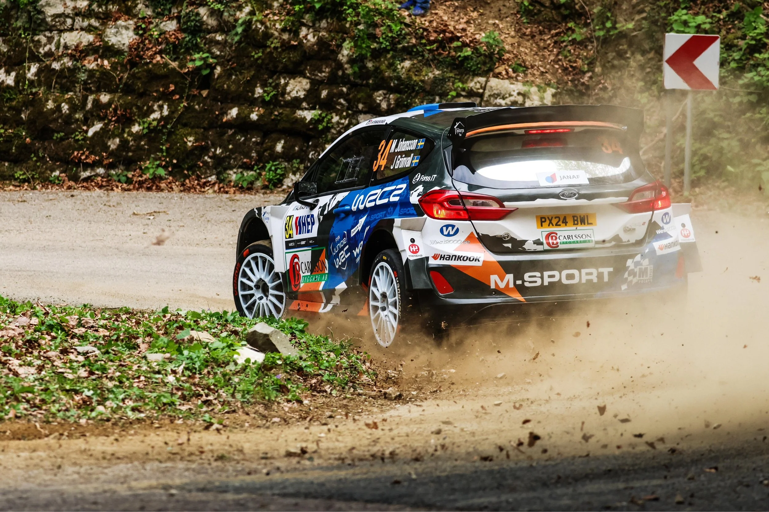 A rally car with the number 34 is kicking up dust as it navigates a dirt rally stage on a forested course, with a red and white arrow sign indicating a sharp turn ahead.